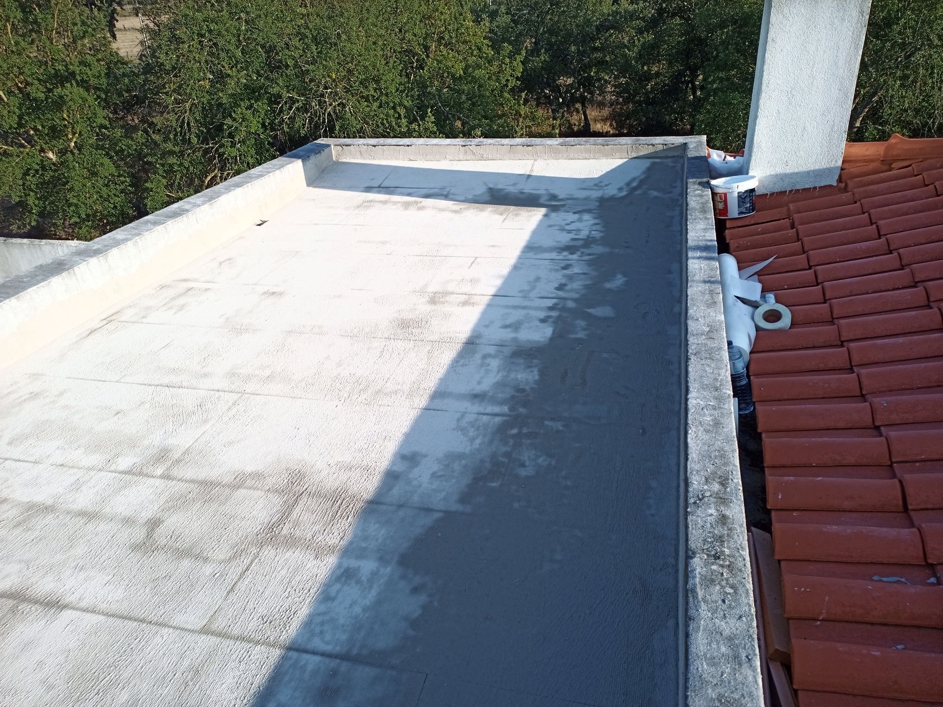 Telhado plano com revestimento cinza ao lado de um telhado de telhas vermelhas e árvores ao fundo.