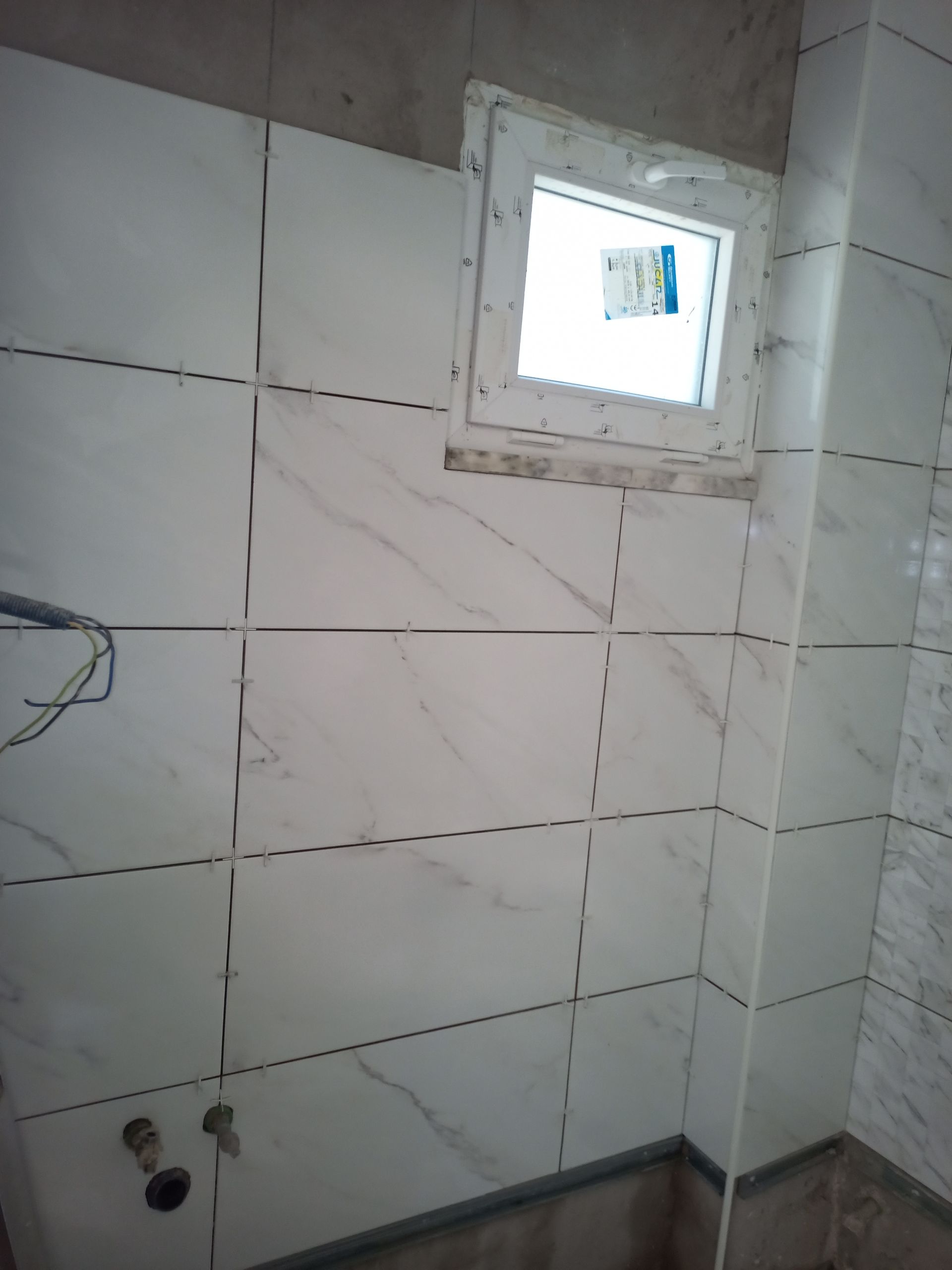 Banheiro com azulejos brancos imitando mármore, uma pequena janela e parede de gesso inacabada.