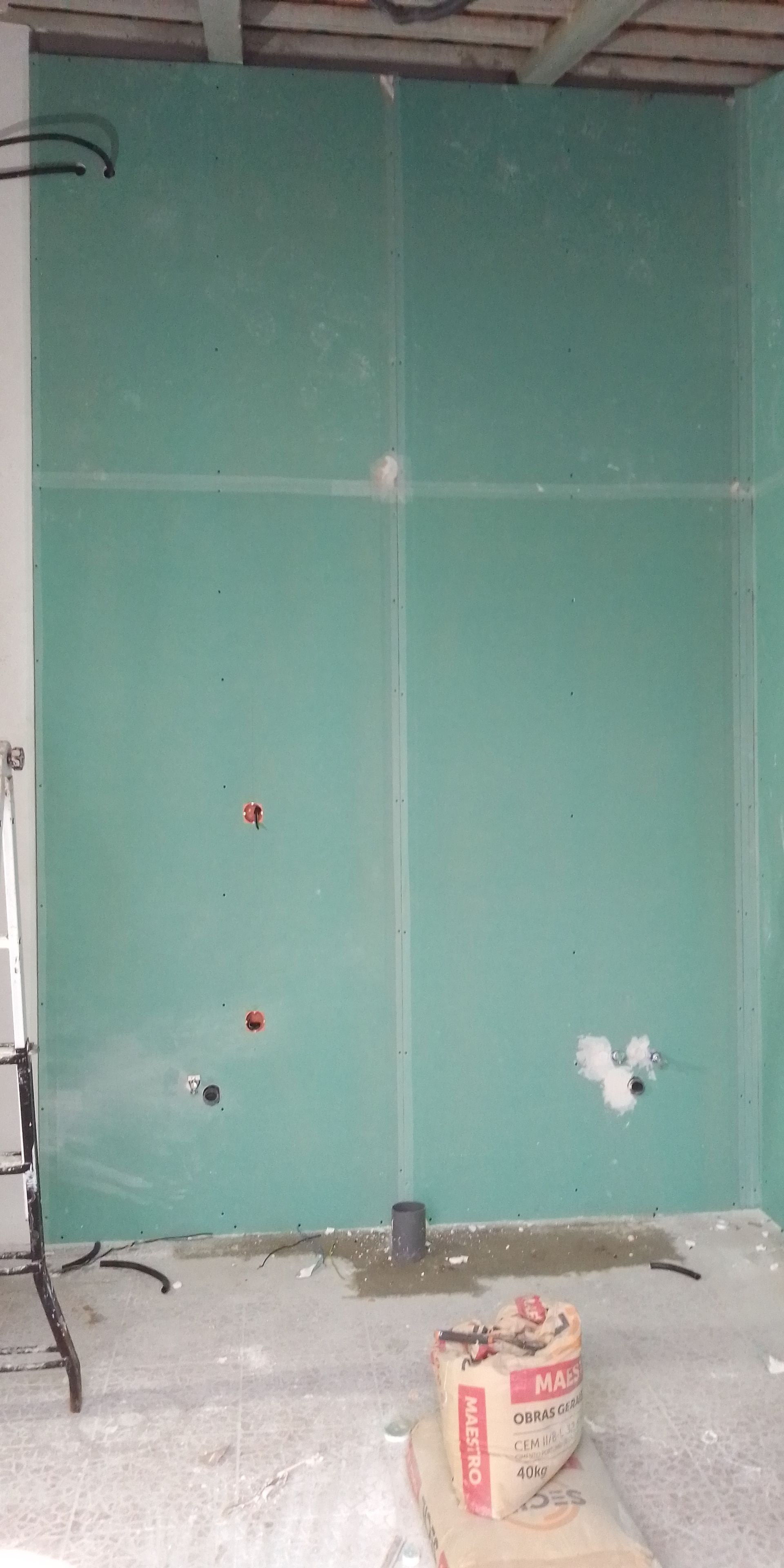Parede interna em construção com drywall verde, instalações elétricas e hidráulicas visíveis e um saco de cimento.