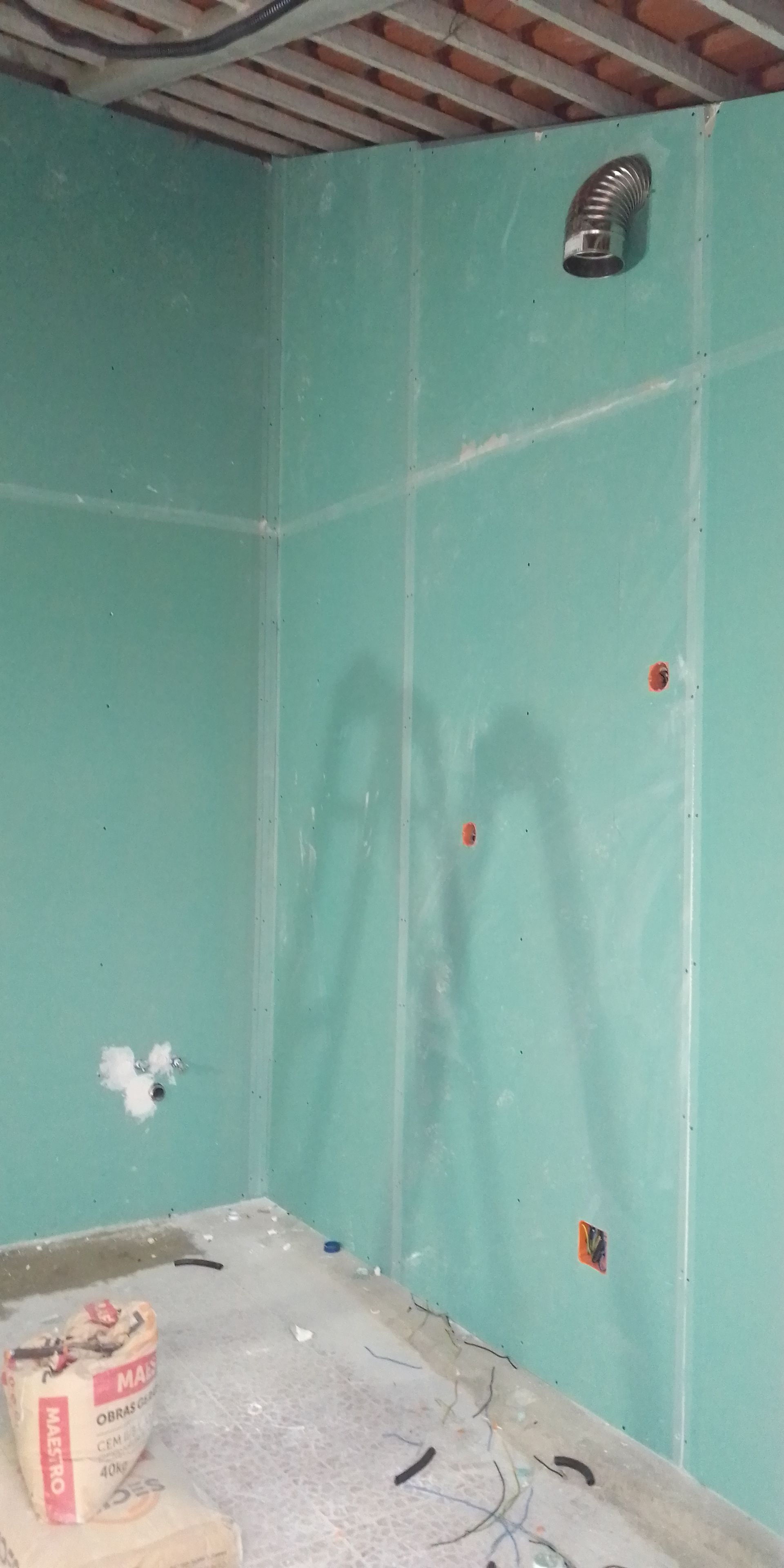 Interior de um cômodo em construção com paredes de gesso acartonado verde. Tomadas elétricas e tubulações são visíveis.