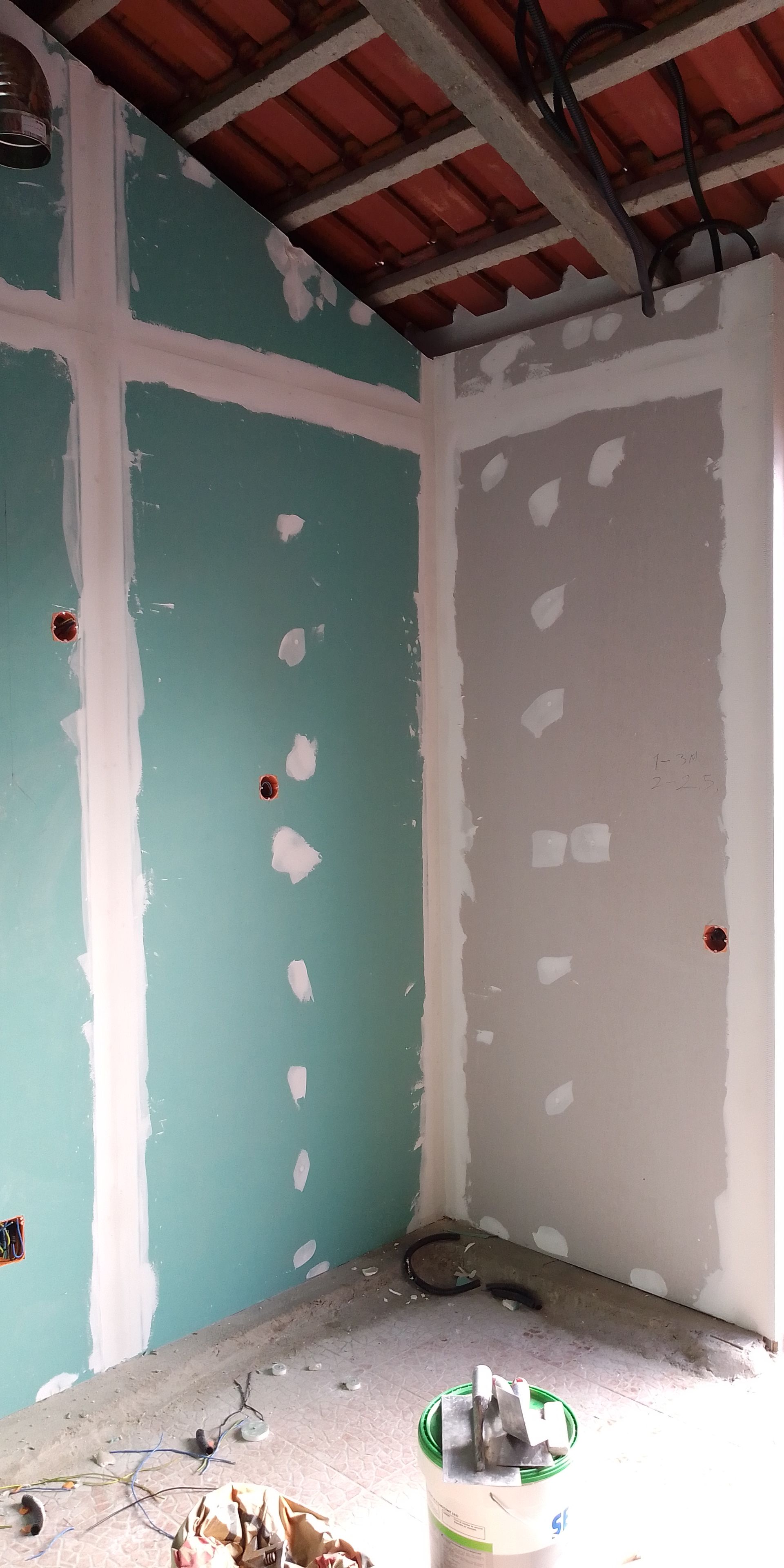 Canto interno de um cômodo com instalação de drywall em andamento; as paredes são em tons de verde-azulado e cinza; o teto é de madeira aparente.