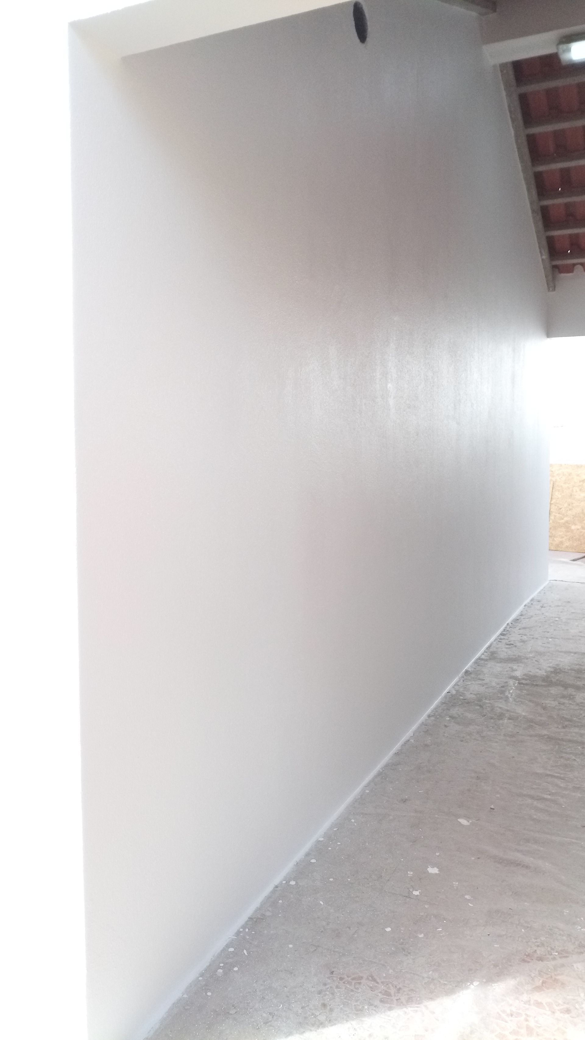 Parede pintada de branco ao longo de uma passarela externa, com vista parcial para um teto de tijolos e piso de concreto.