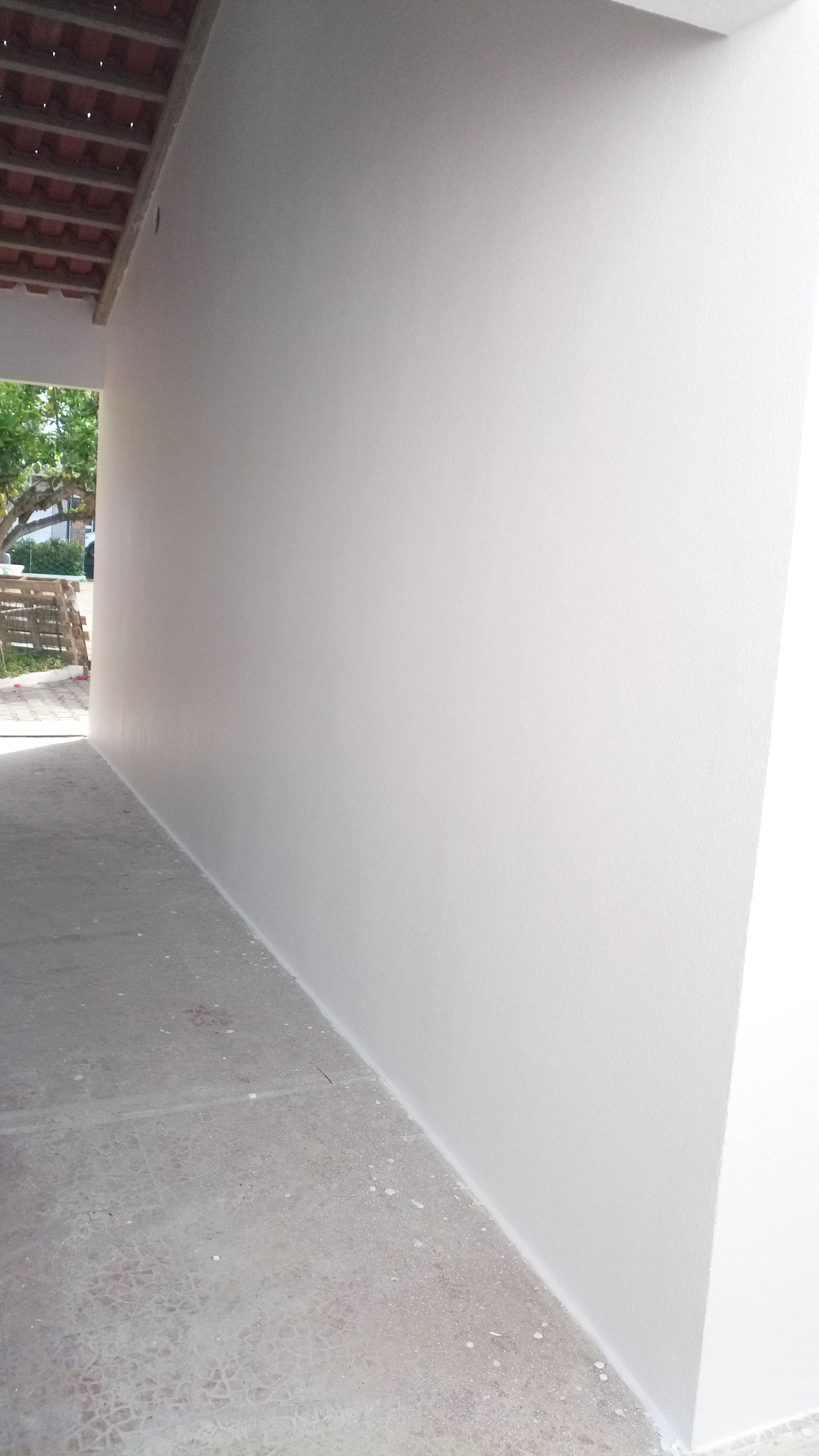 Caminho de concreto cinza ao lado de uma parede pintada de branco, sob uma cobertura de telhas.