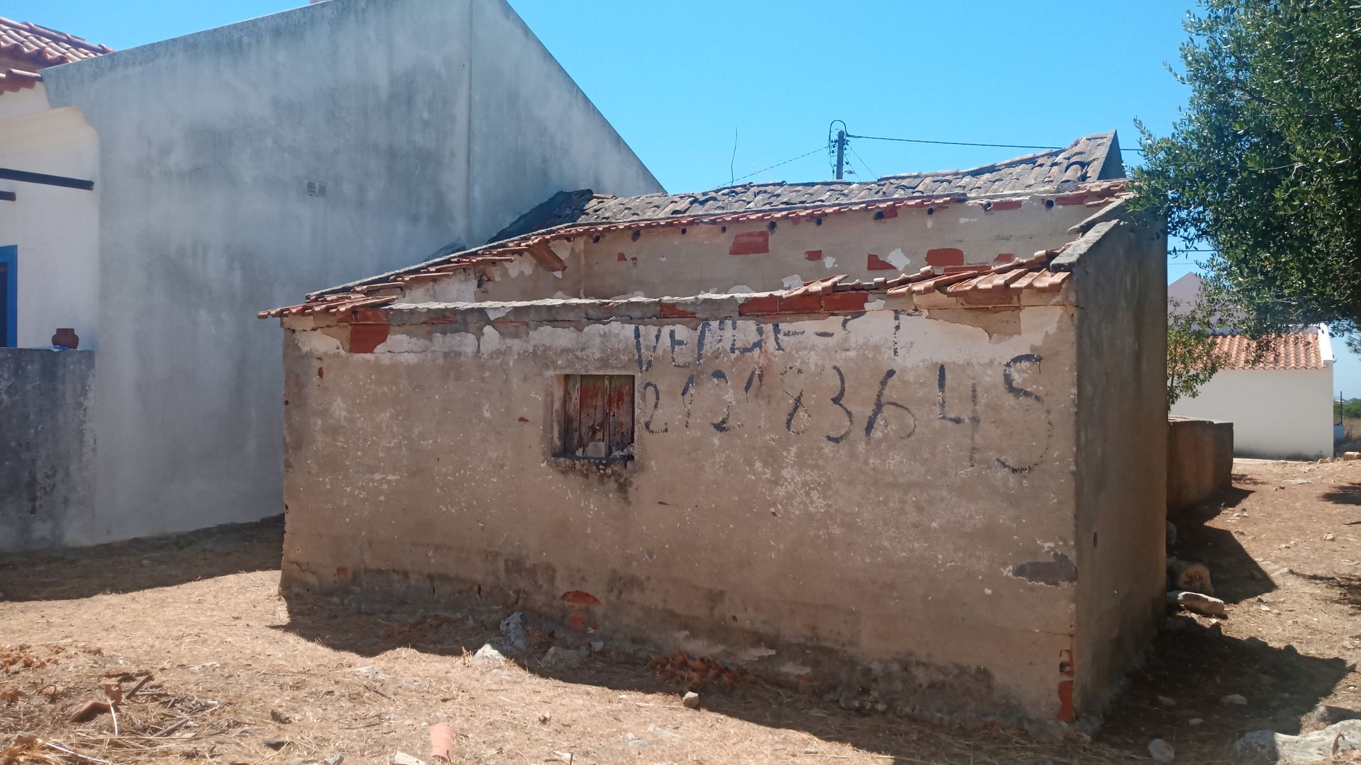 Prédio desgastado pelo tempo, com telhado em ruínas e pichações, situado em uma área rural seca e ensolarada.