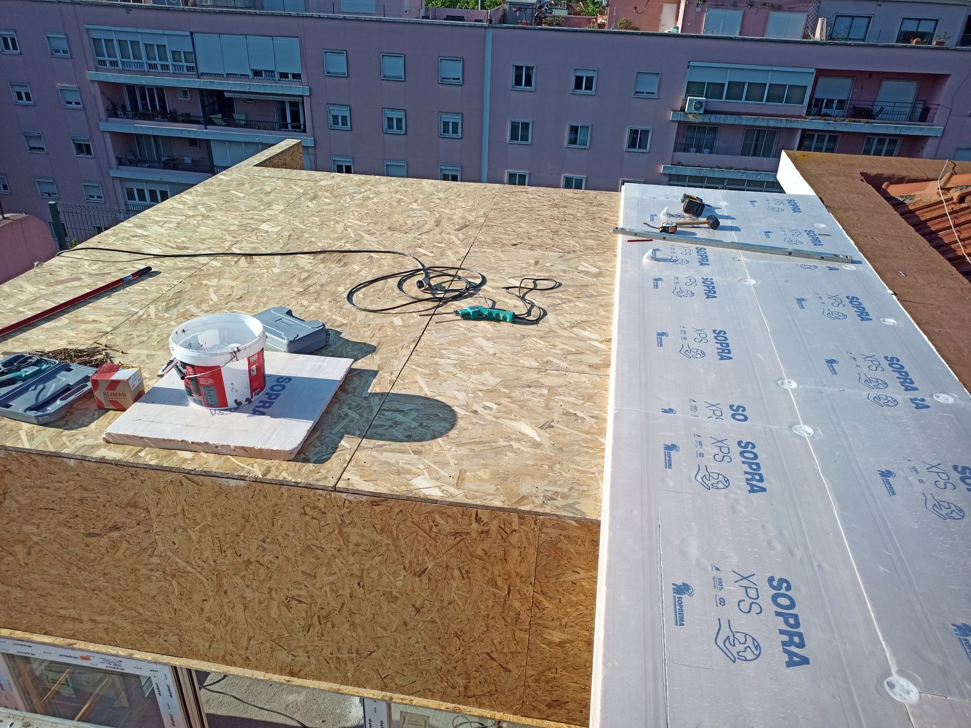 Construção de telhado: placas de OSB, isolamento, ferramentas e materiais. Prédios da cidade ao fundo em um dia ensolarado.