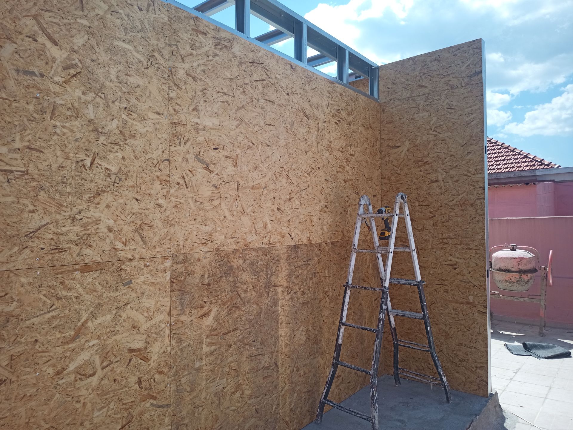 Construção de uma estrutura com painéis de madeira e armação de metal. Uma escada encostada na parede sob um céu azul.