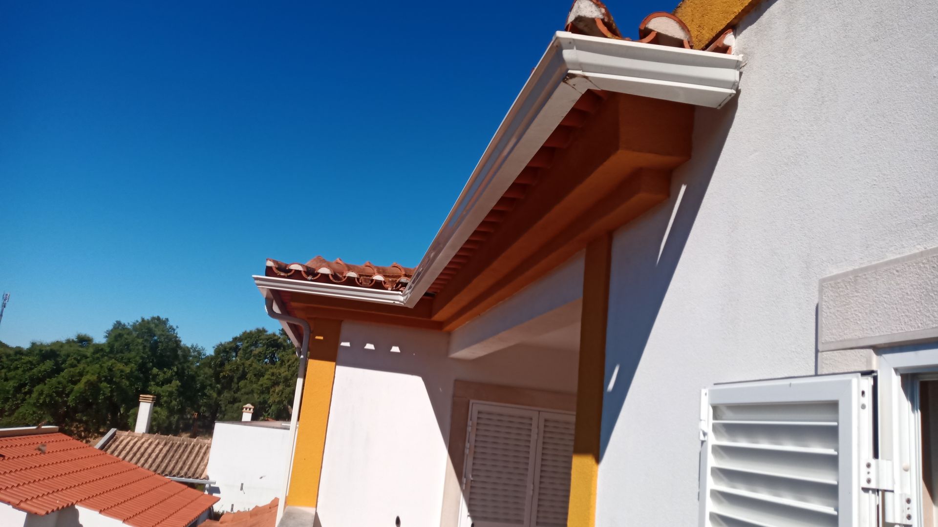 Prédio branco com detalhes e calhas laranja, telhado de telha vermelha e céu azul.