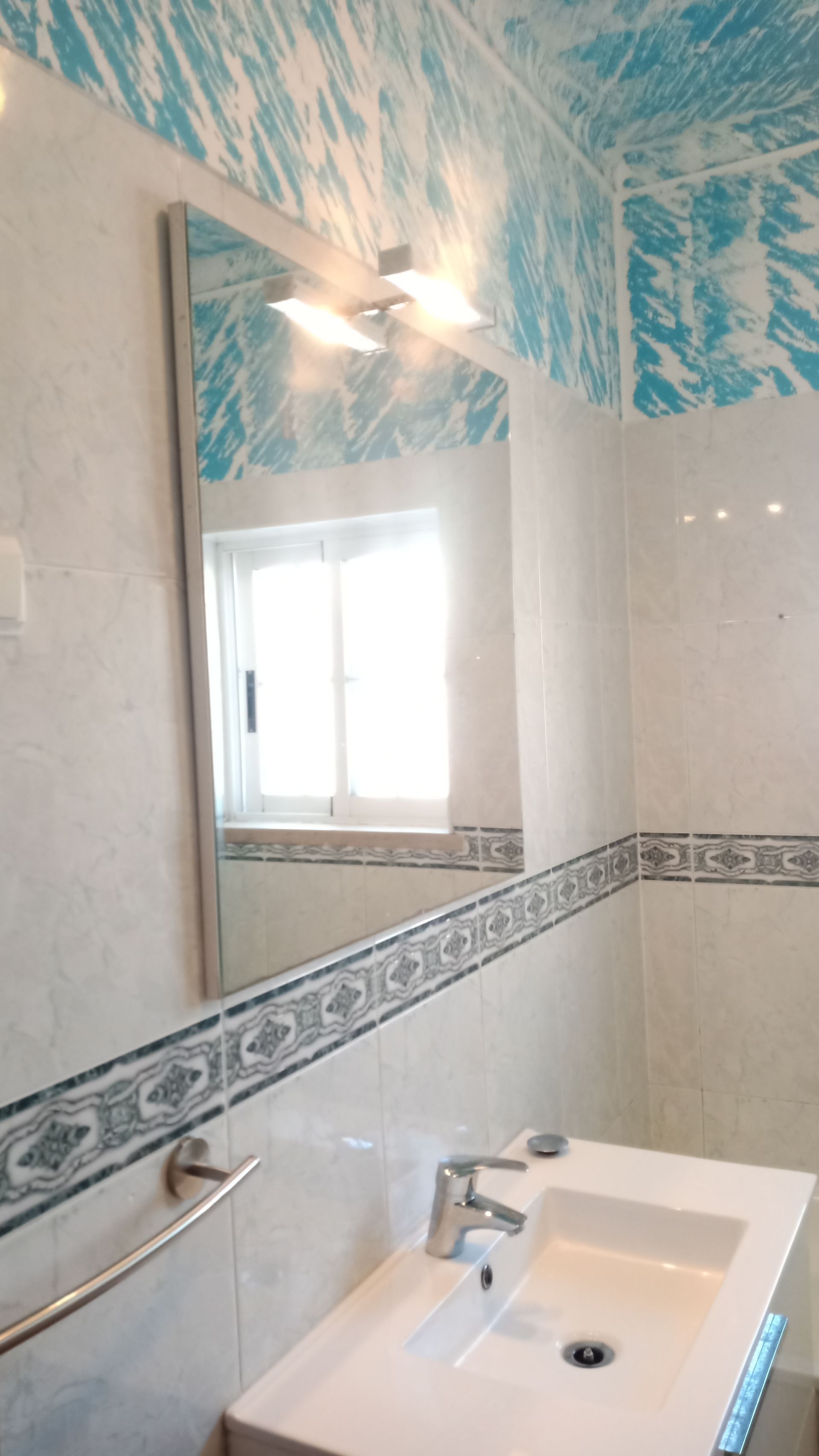 Banheiro com pia branca, espelho e azulejos de parede com estampa azul.