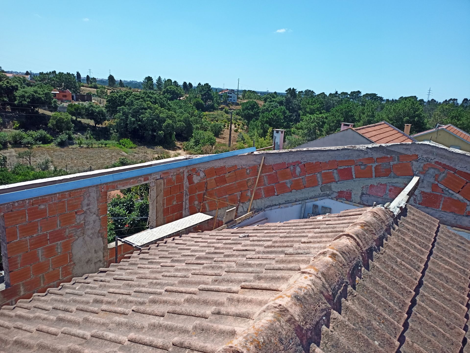 Vista do terraço de um edifício com fachada de tijolos, telhas de barro e árvores verdes ao redor, sob um céu azul.