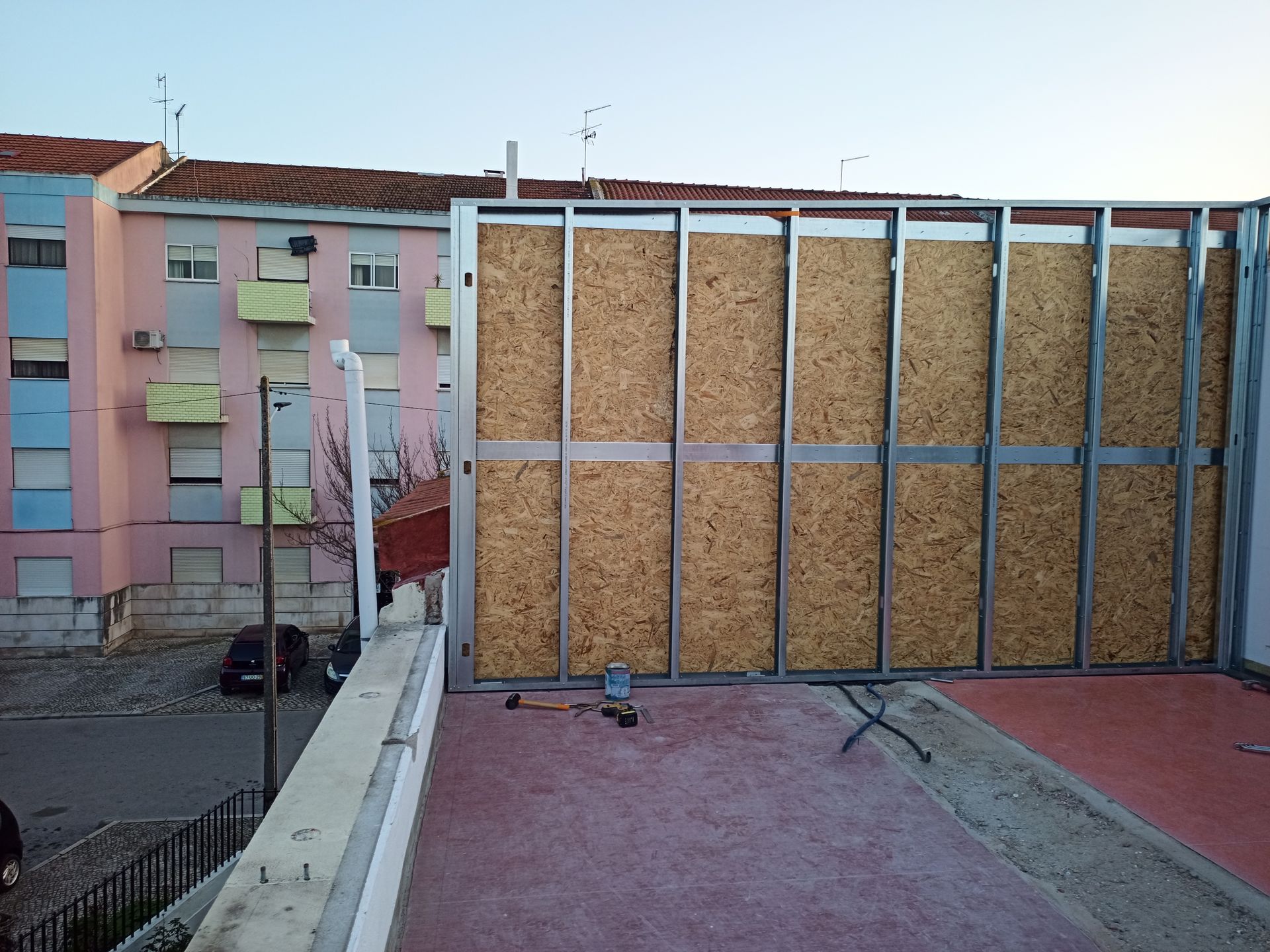Parede com estrutura metálica e painéis de cortiça sendo construída em um telhado, com prédios residenciais ao fundo.
