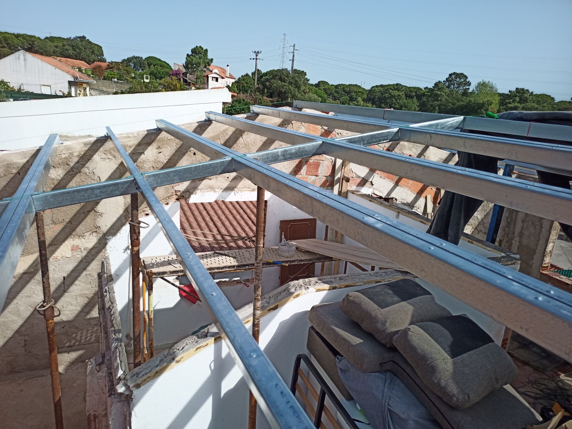 Telhado em construção com vigas metálicas, luz solar e paisagem urbana ao fundo.