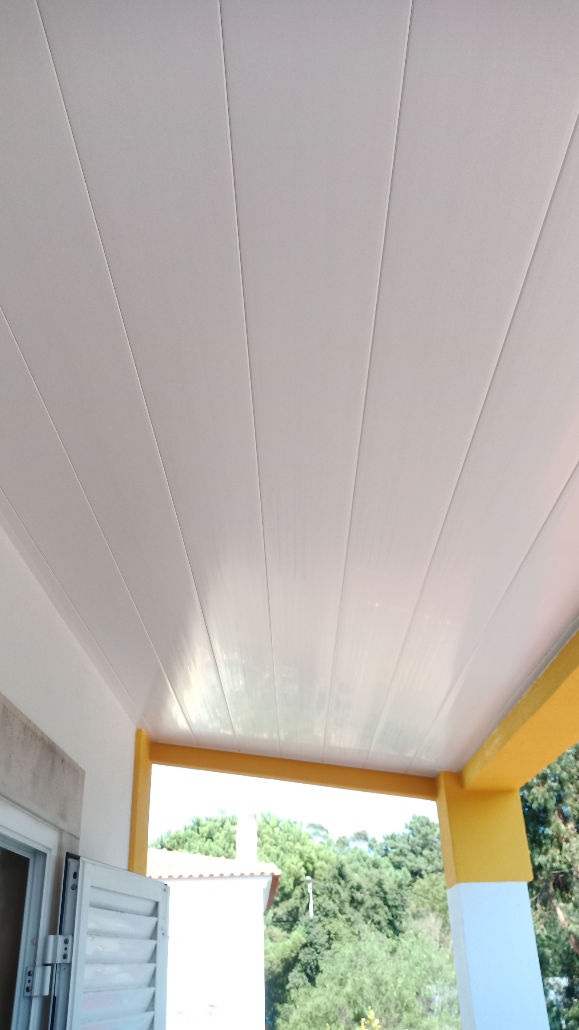 Teto branco com linhas ranhuradas, parte de uma varanda. Pilares de sustentação amarelos e brancos. Árvores verdes ao fundo.