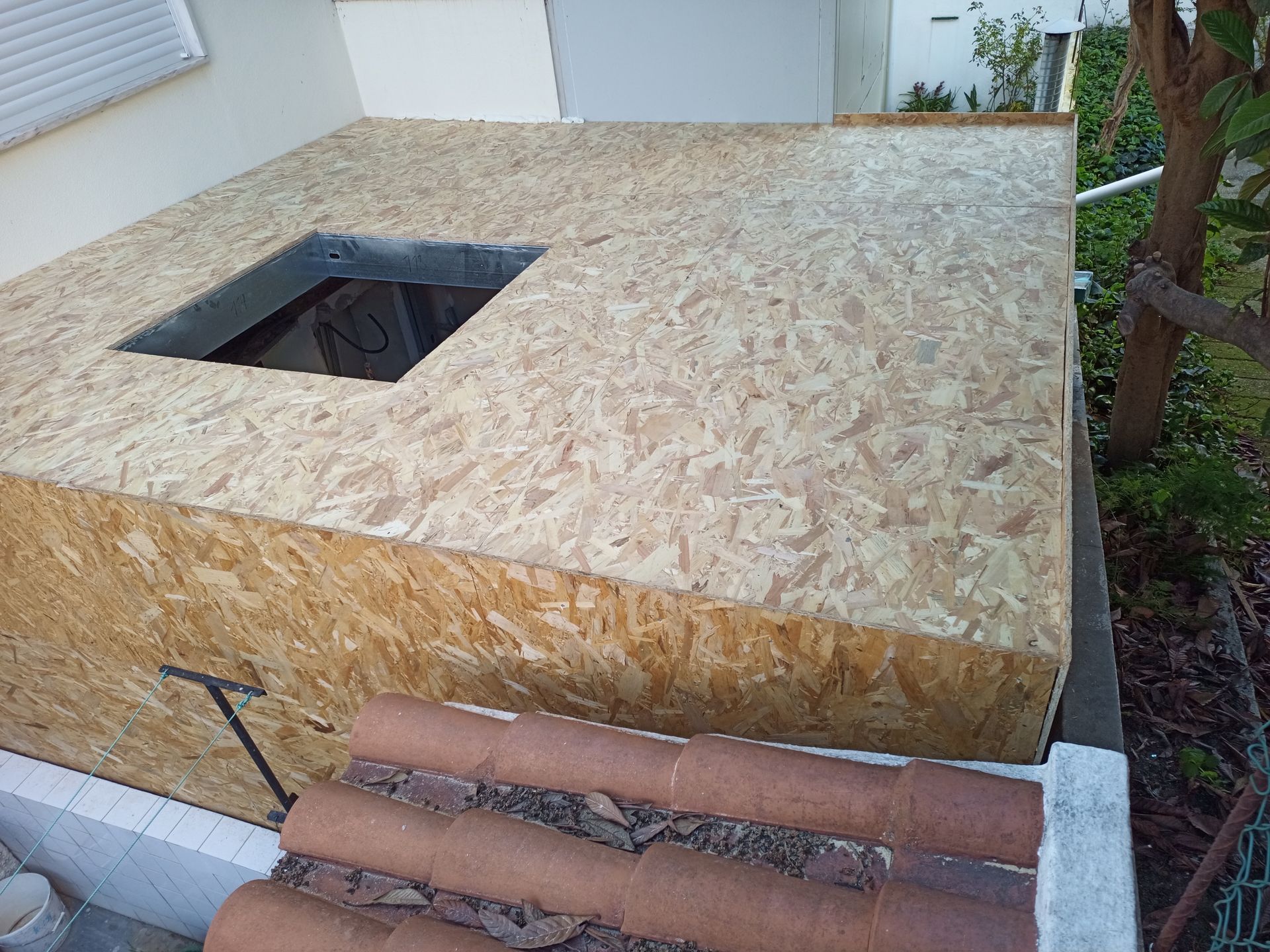 Uma estrutura de madeira com uma abertura quadrada. Telhas marrons são visíveis em primeiro plano, com vegetação ao fundo.