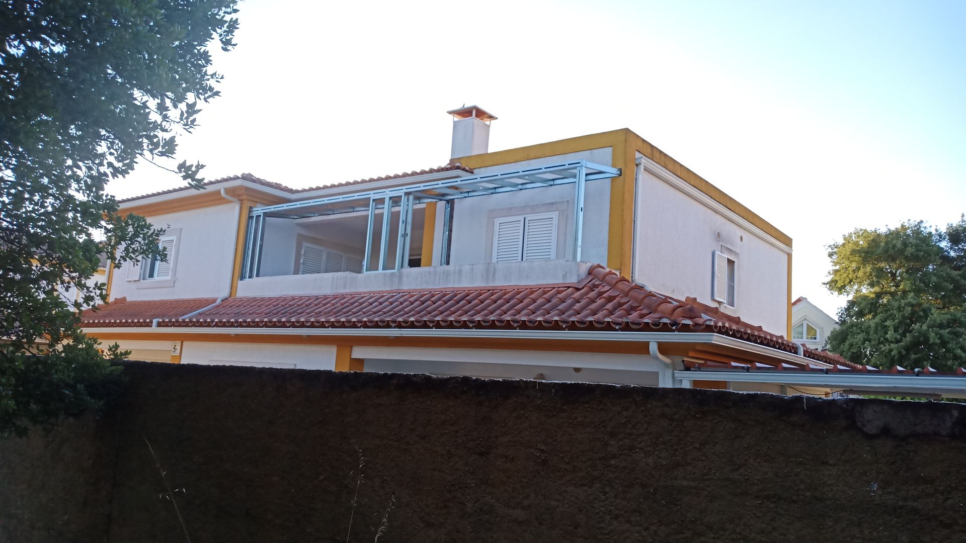 Casa de dois andares com telhado de telha vermelha, paredes brancas com detalhes em amarelo e varanda com estrutura metálica; situada ao ar livre.