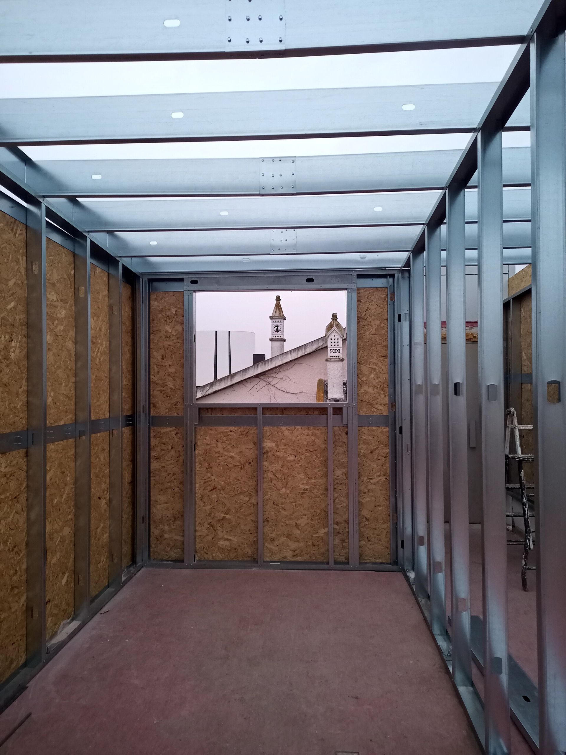 Vista interior de uma sala com estrutura metálica em construção, com paredes de aglomerado expostas e uma janela com vista para os telhados.