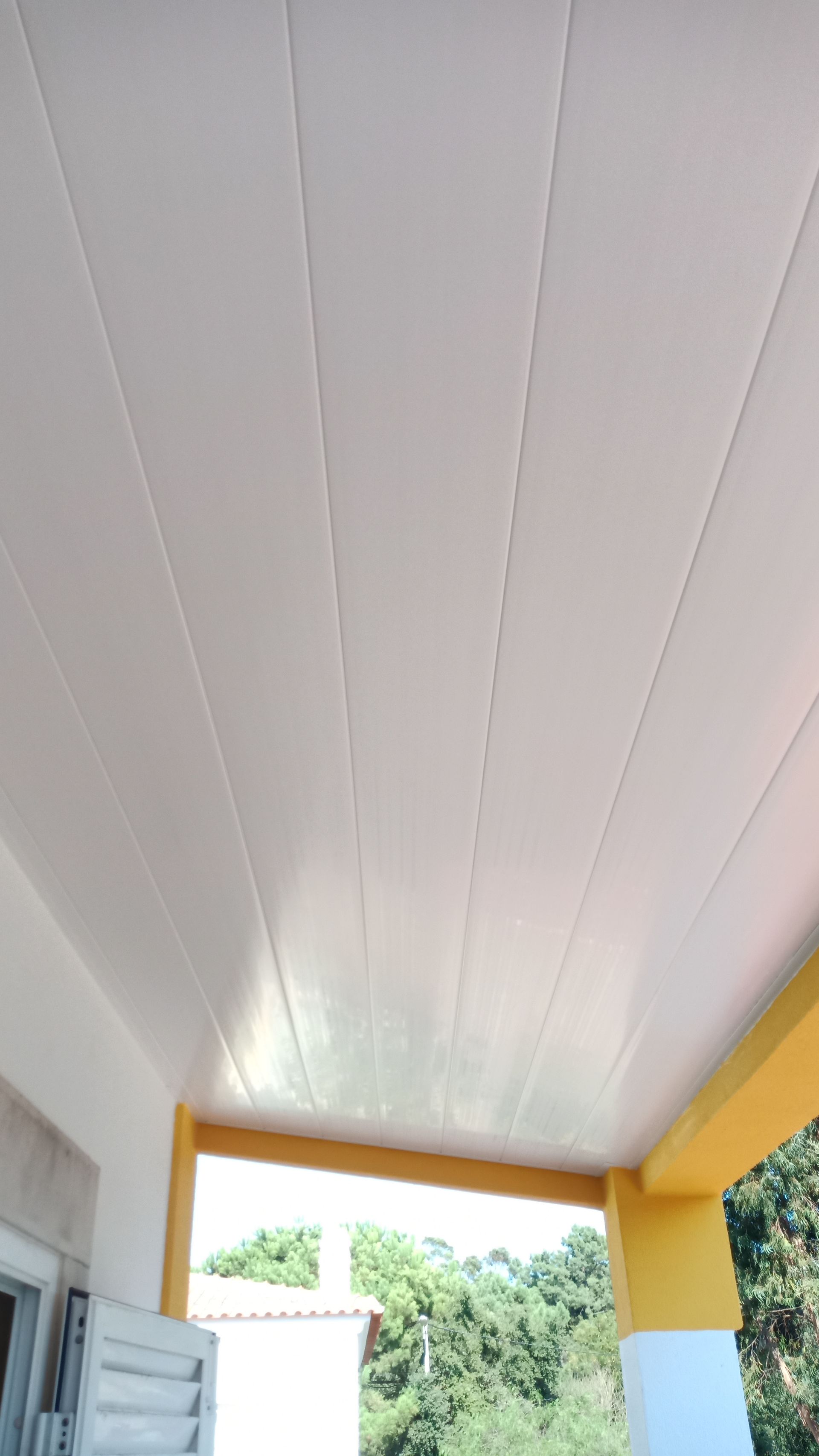 Teto com painéis brancos em uma varanda com colunas de sustentação amarelas; árvores verdejantes ao fundo.