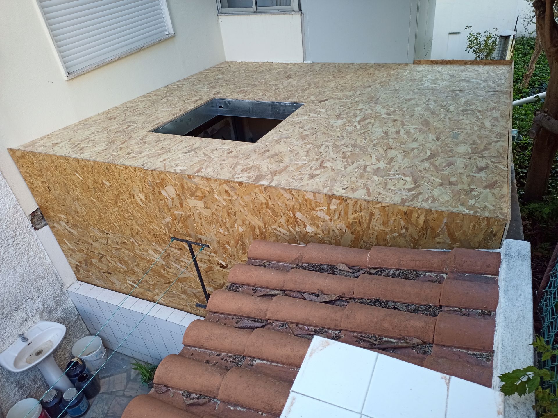 Uma estrutura em forma de caixa feita de aglomerado de madeira com uma abertura quadrada no topo, assente sobre um telhado de telhas.