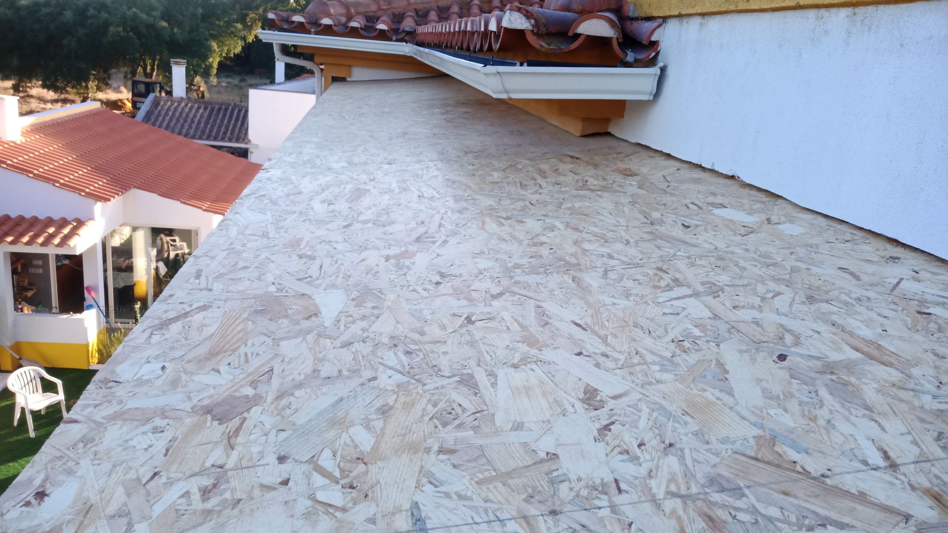 Telhado de madeira OSB em construção próximo a uma casa com telhado de telha vermelha e paredes brancas.
