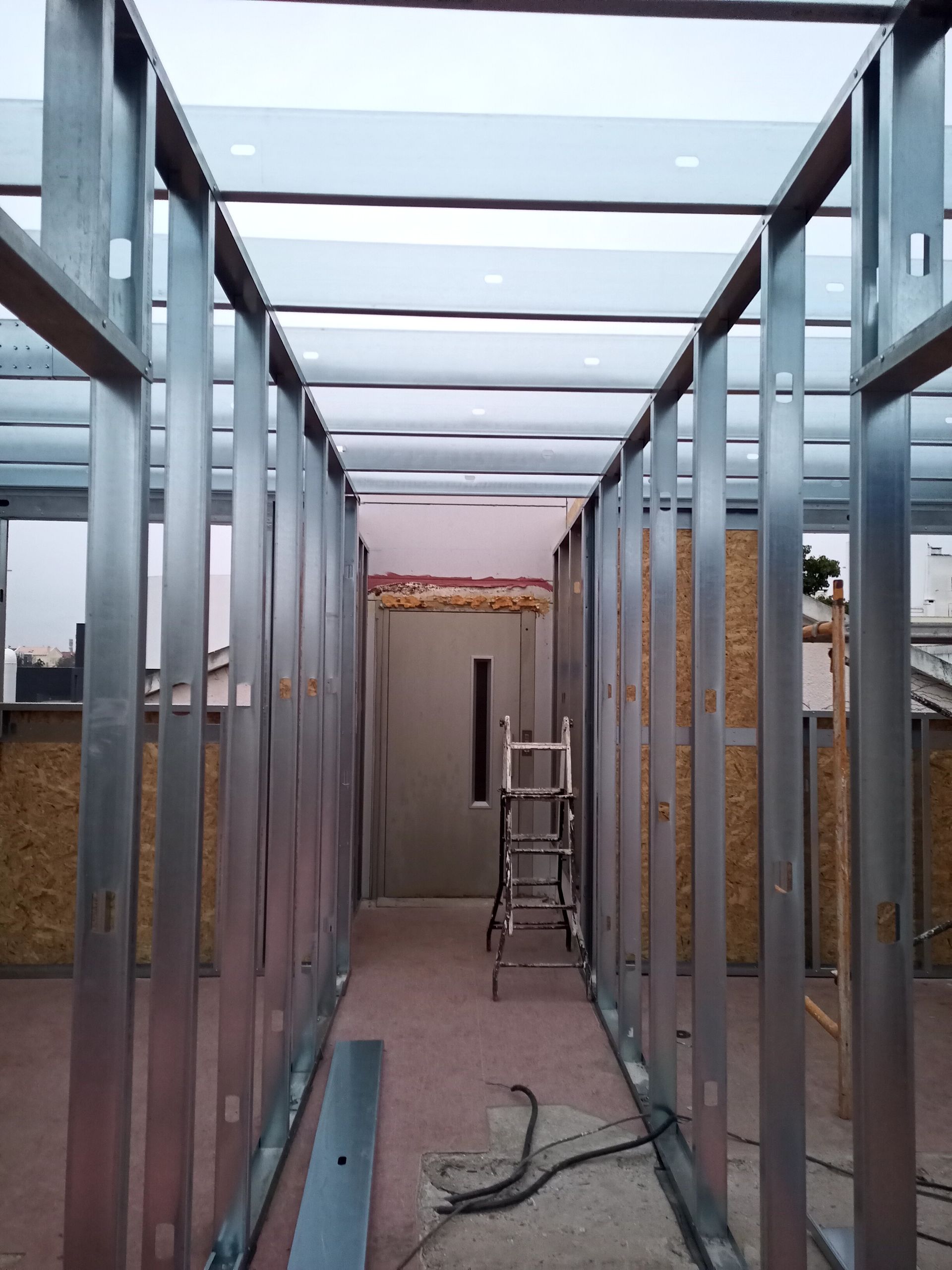 Estrutura metálica de um corredor em construção, com uma escada e uma porta visíveis ao fundo.