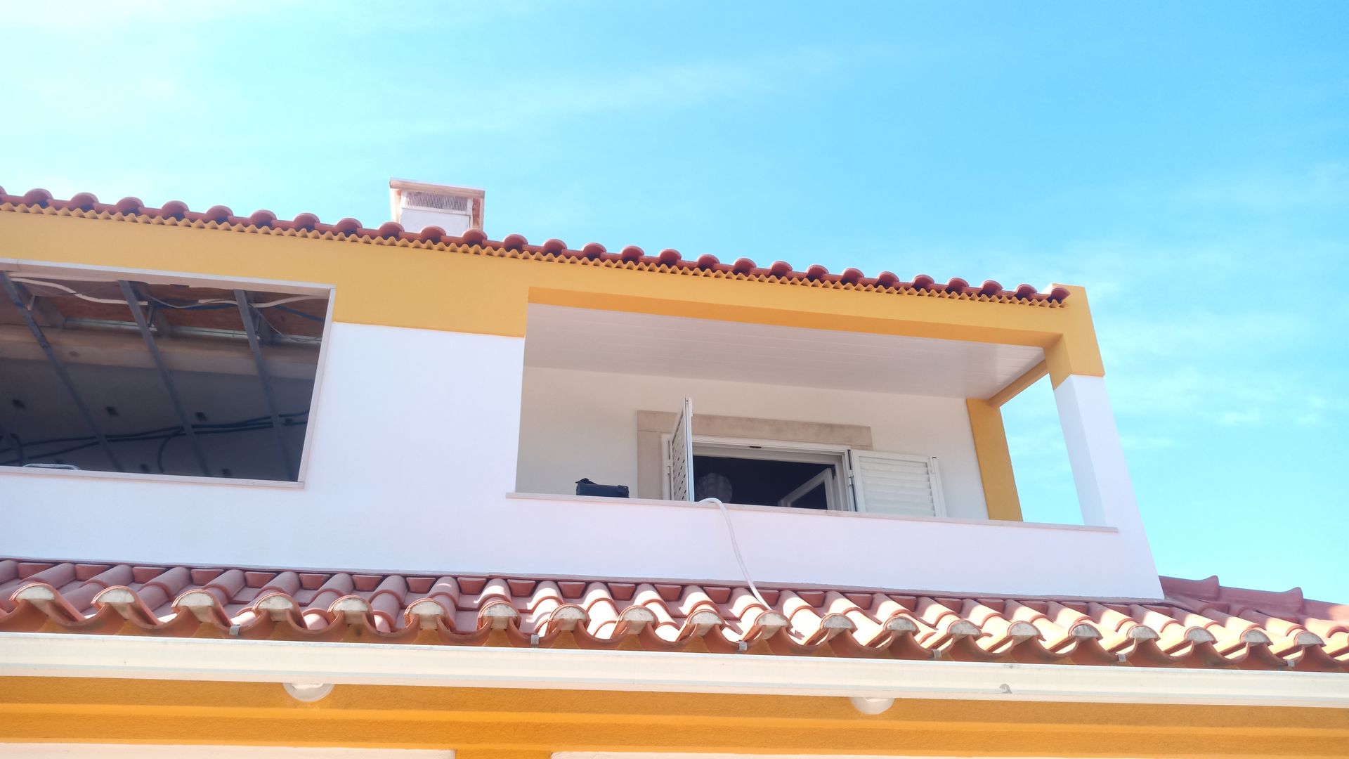 Prédio de dois andares com detalhes em amarelo, paredes brancas e telhado de telhas, sob um céu azul brilhante.