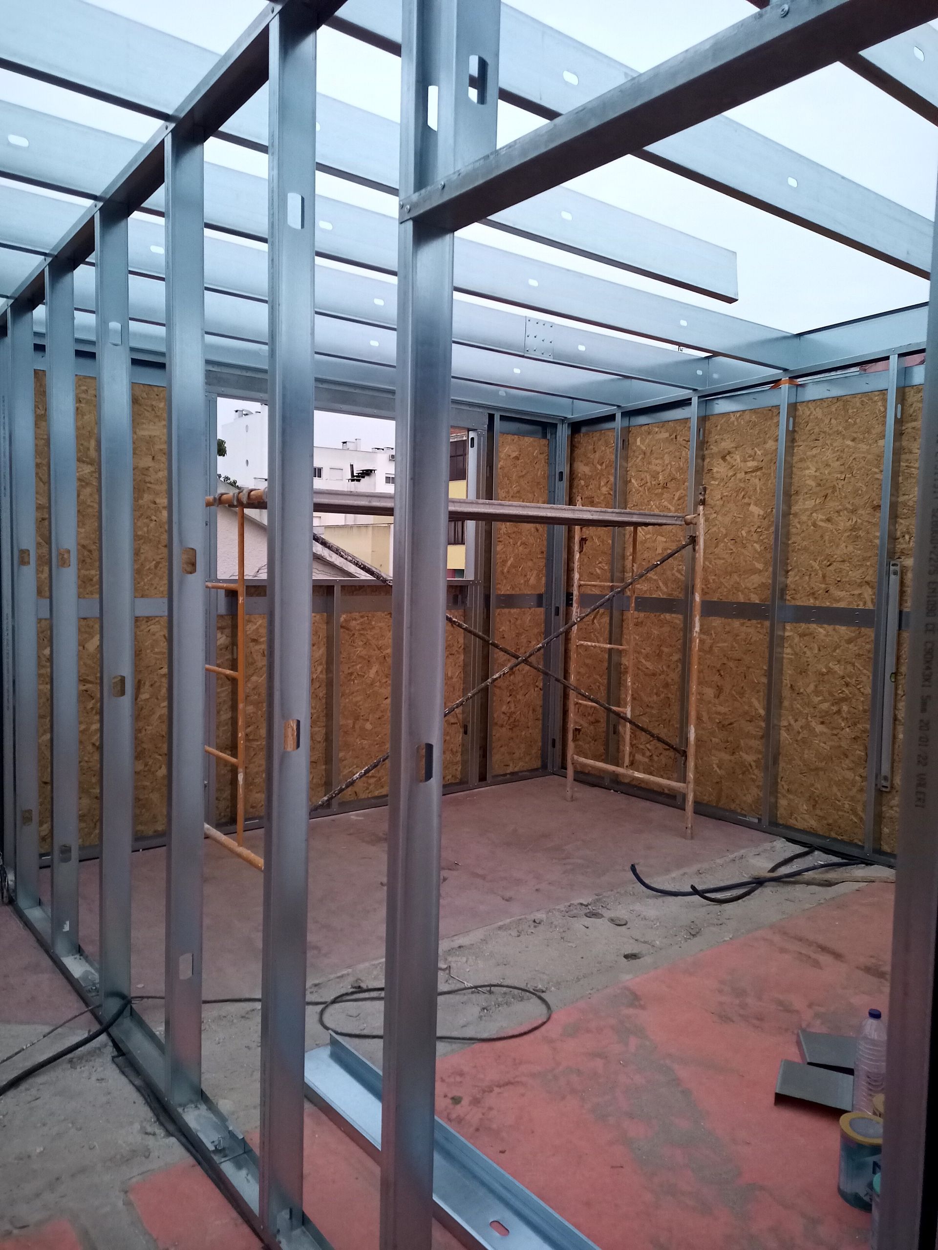 Estrutura metálica de um edifício em construção, com painéis de madeira nas paredes. Andaimes visíveis.