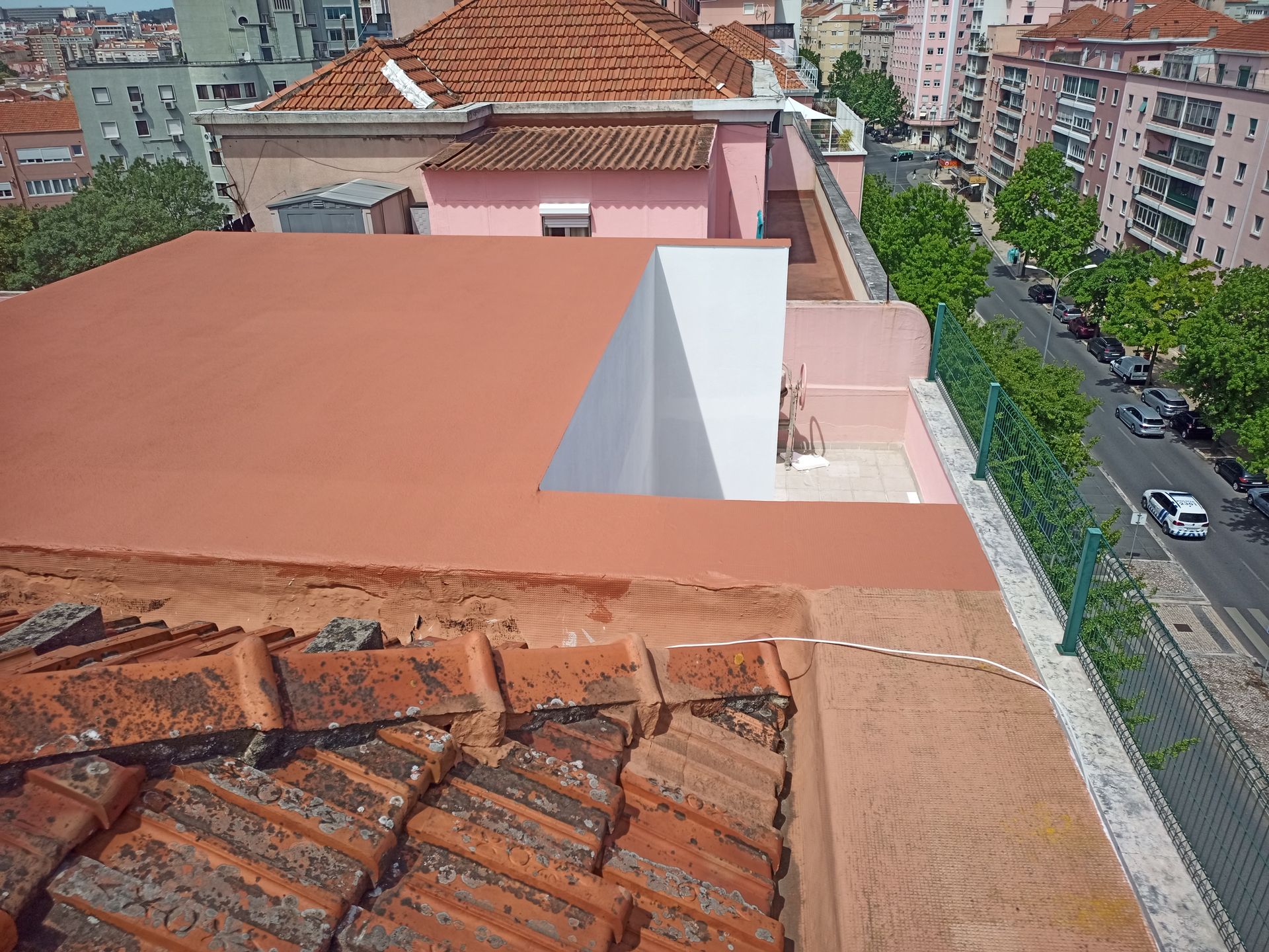 Borda do telhado de telhas vermelhas com cobertura plana e vermelha, paredes brancas e vista para a rua com prédios e uma cerca verde.