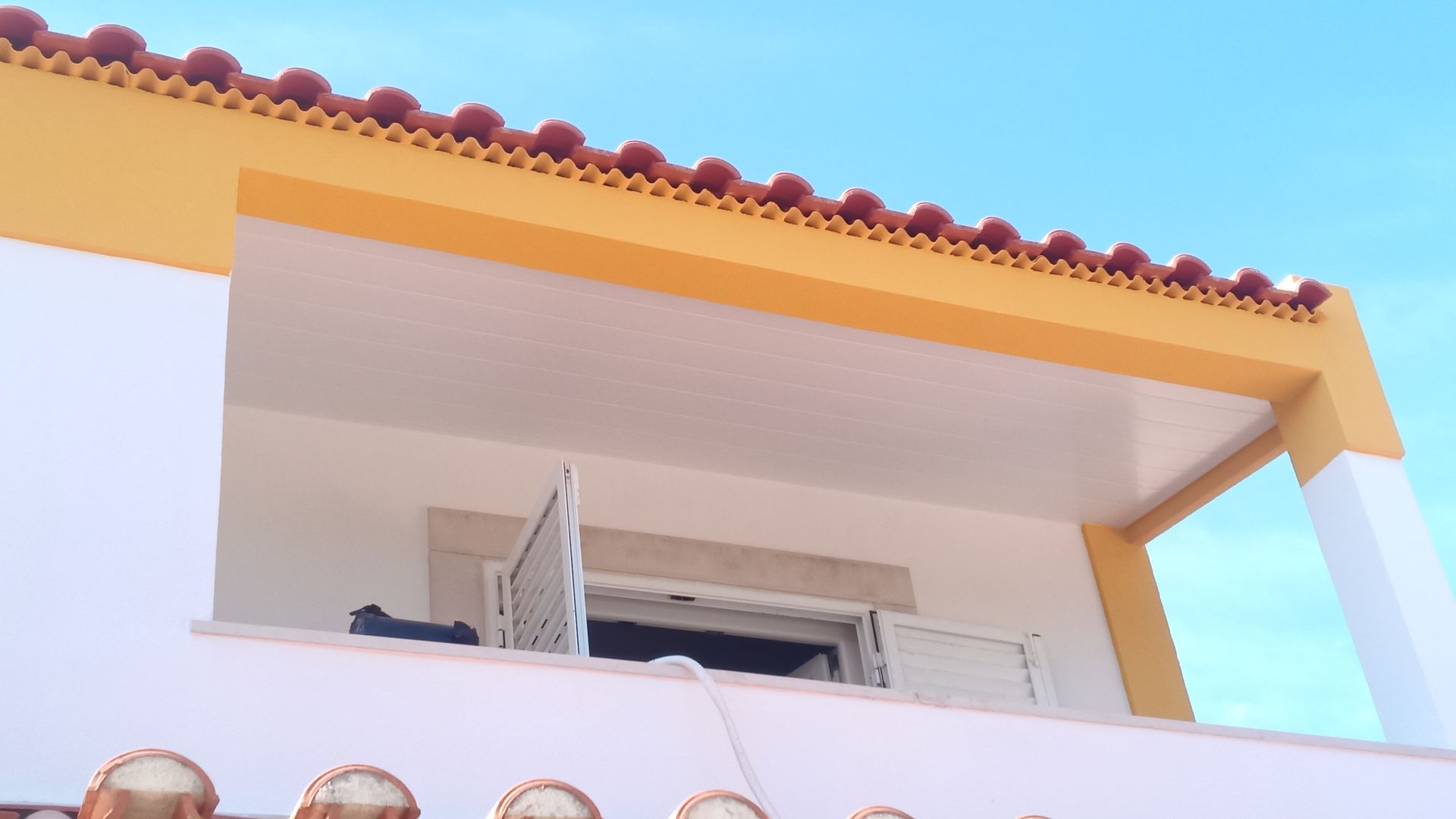 Prédio branco e amarelo com varanda; telhado de telha vermelha e céu azul visível.