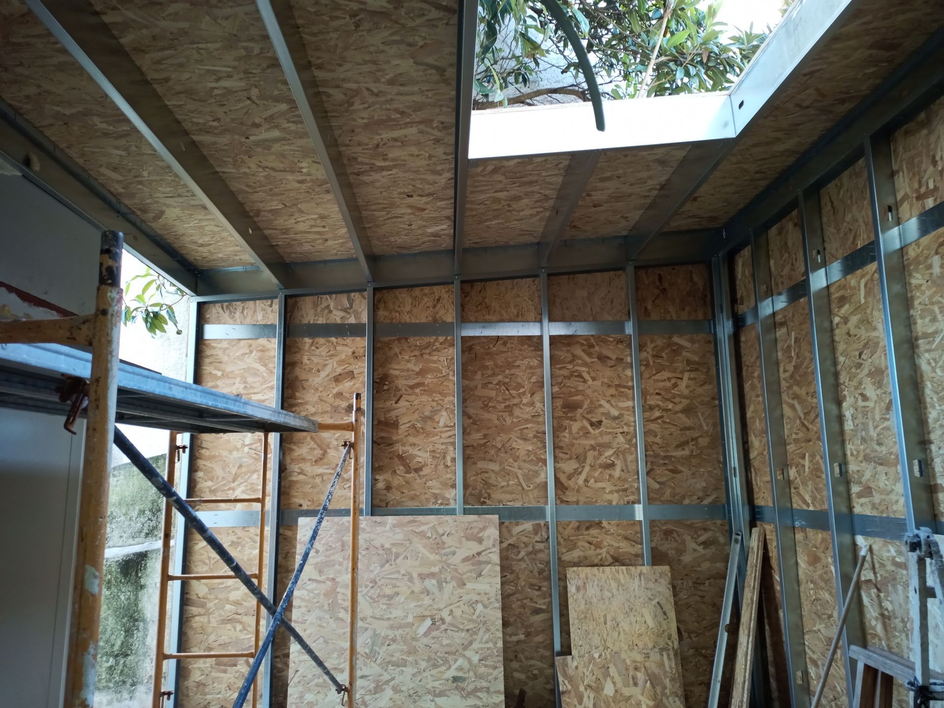 Vista interna de um cômodo em construção, com estrutura metálica e revestimento em OSB. Uma pequena claraboia é visível.