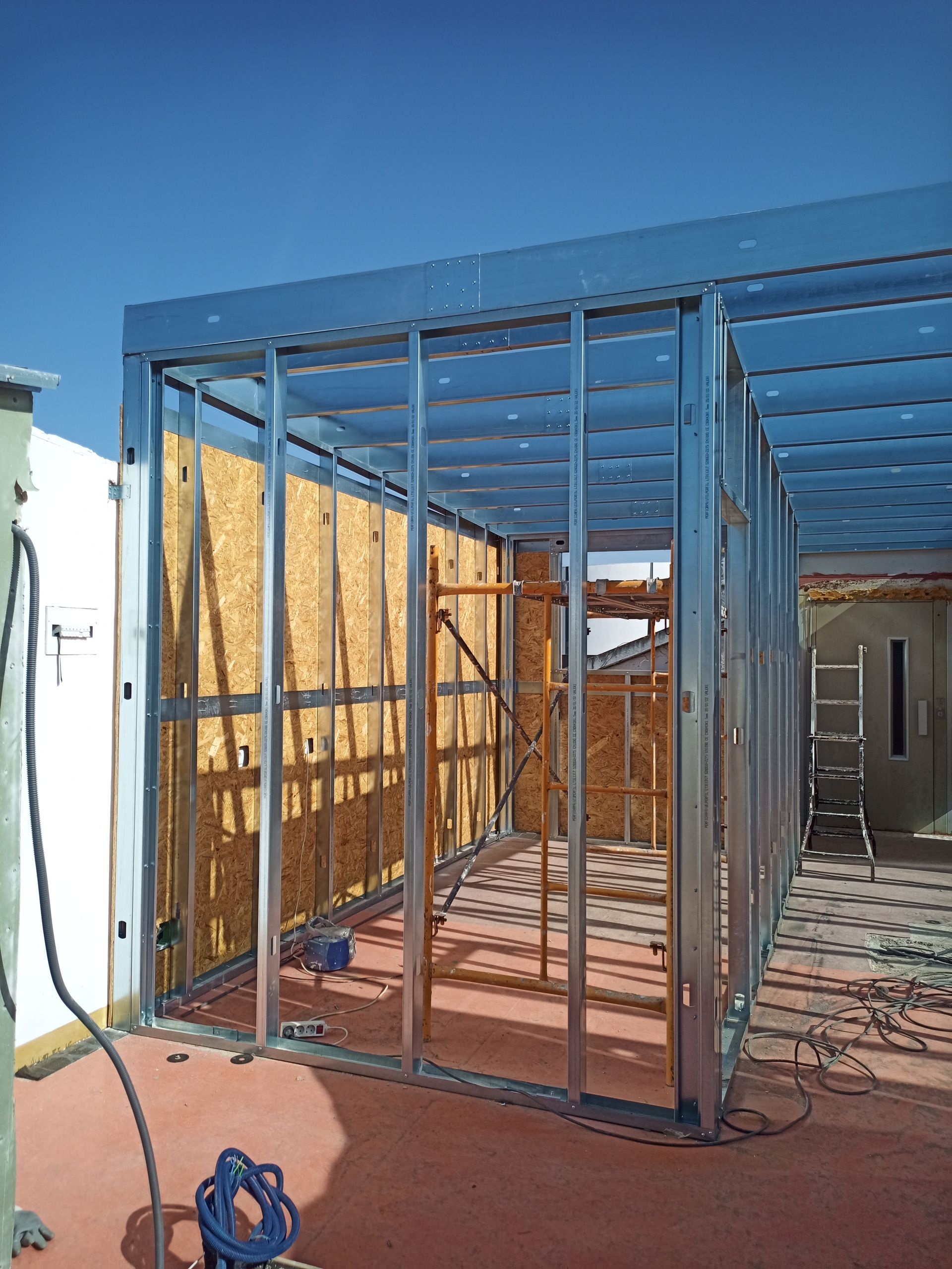 Estrutura metálica de um edifício em construção. As paredes têm revestimento de madeira. Vista do telhado, céu azul.