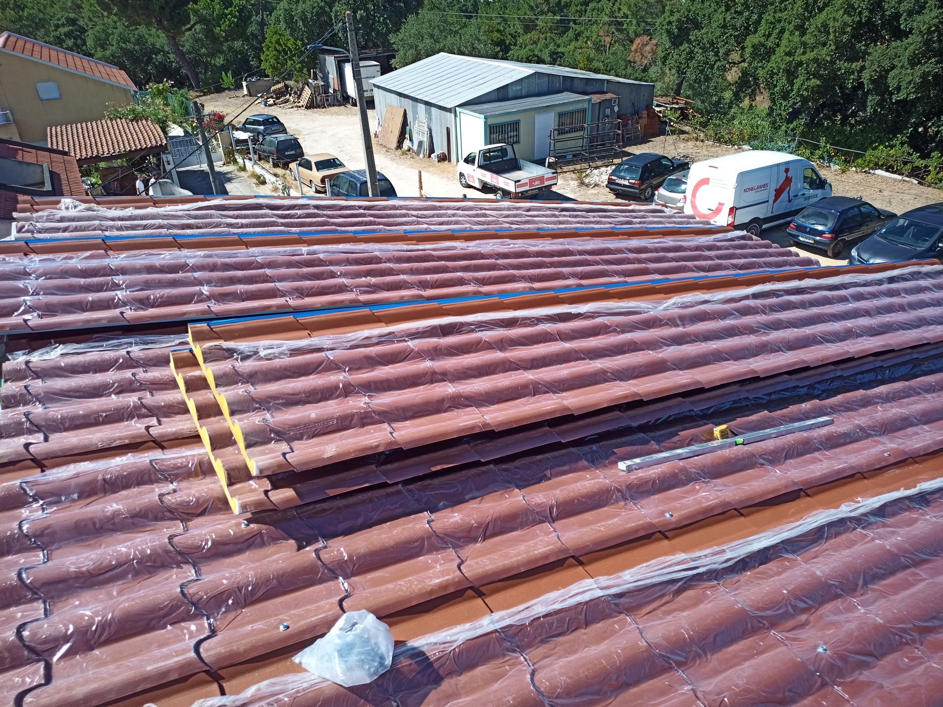 Telhado vermelho ondulado com painéis soltos; veículos e edifícios ao fundo.