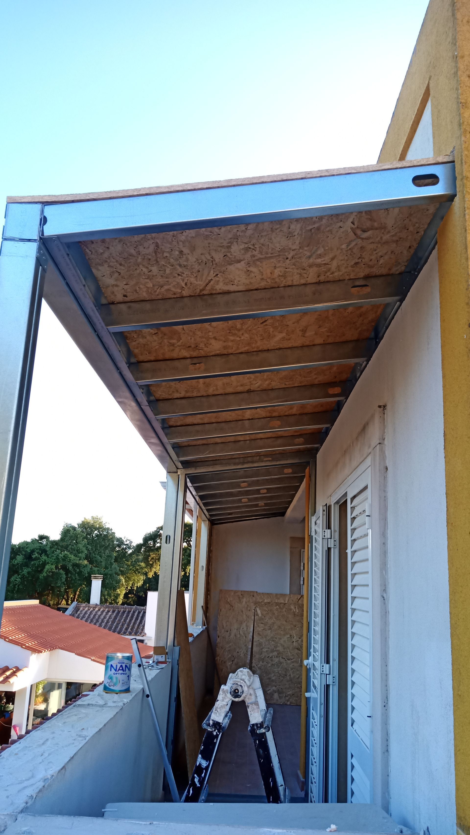Varanda com toldo parcialmente construído em metal e madeira. O céu está azul.
