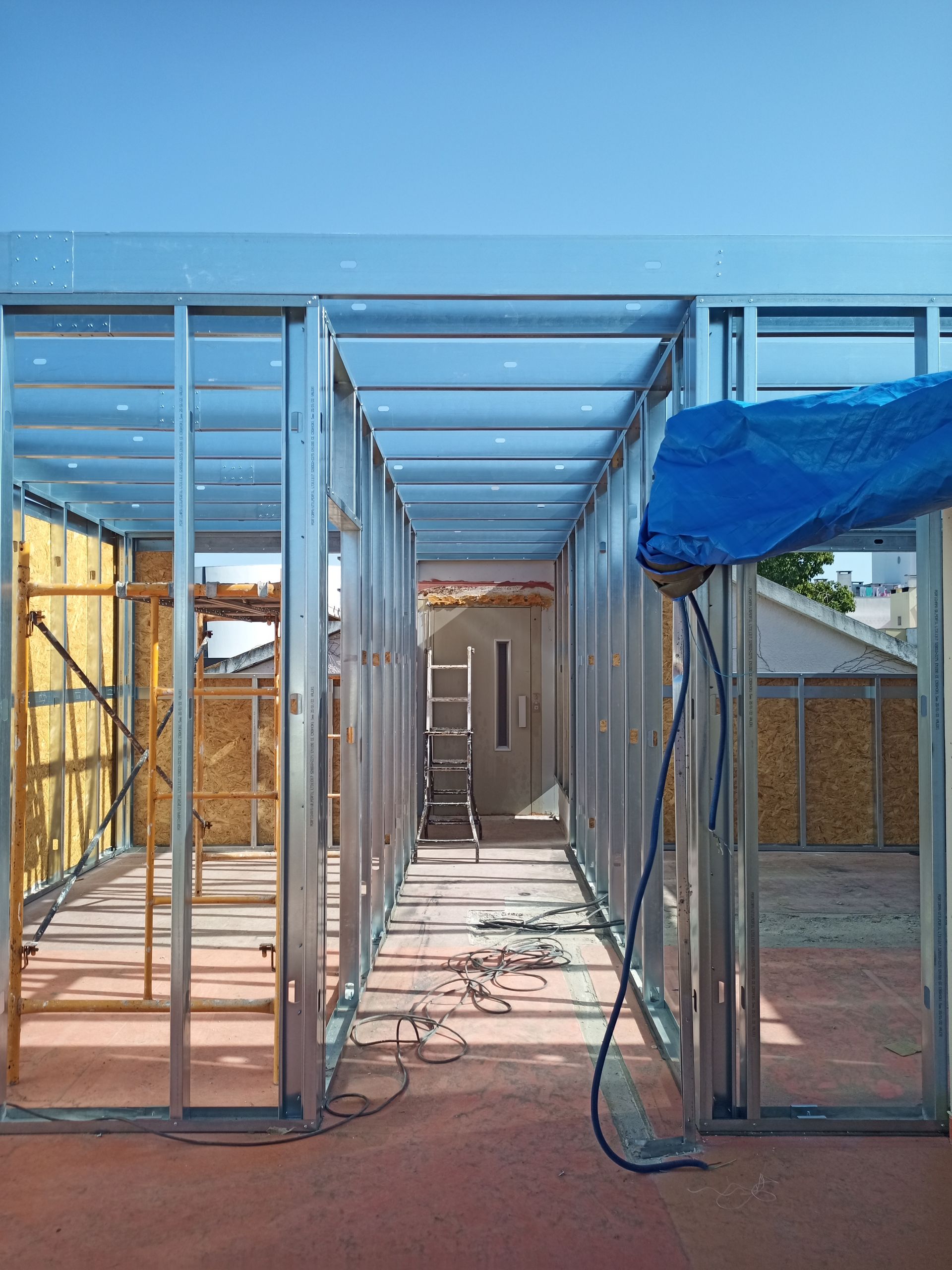 Estrutura metálica de um edifício em construção, com uma lona azul e isolamento visíveis.