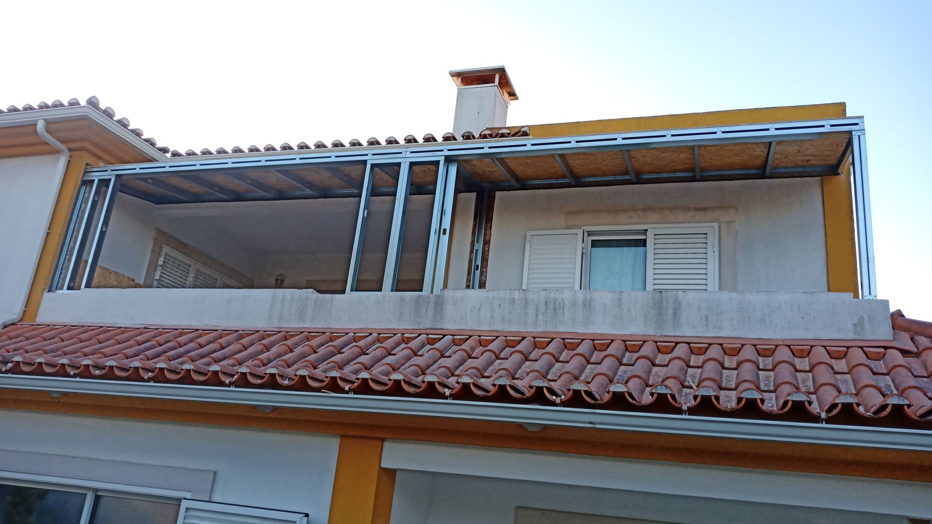 Construção da estrutura de uma varanda externa em uma casa com telhado de telha vermelha e chaminé.