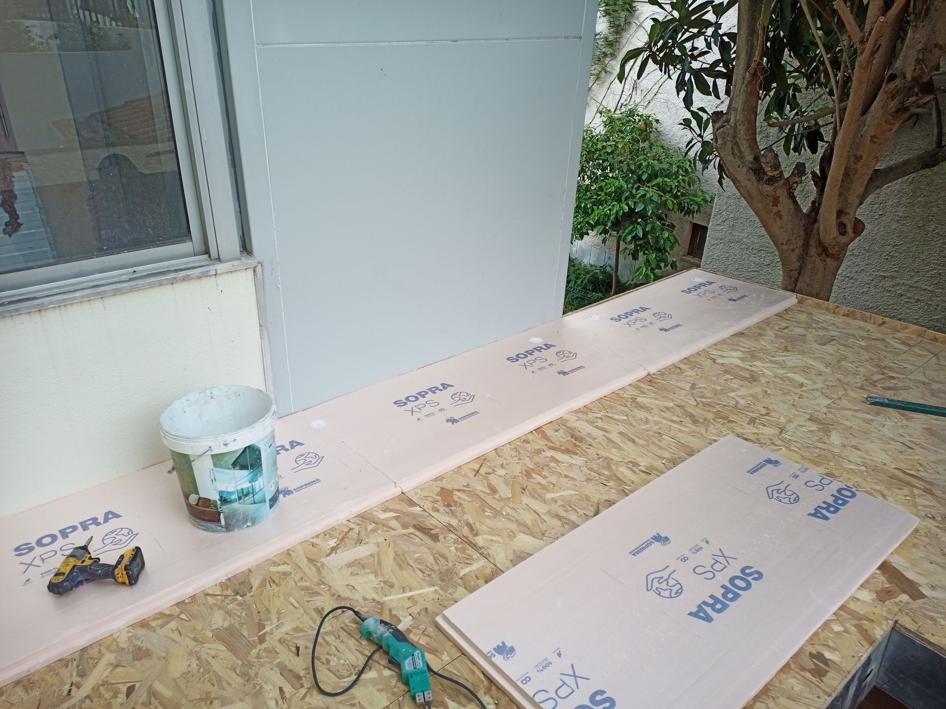 Cena de construção com placas de isolamento rosa sobre uma superfície de madeira compensada. Um balde branco e uma ferramenta elétrica estão presentes.