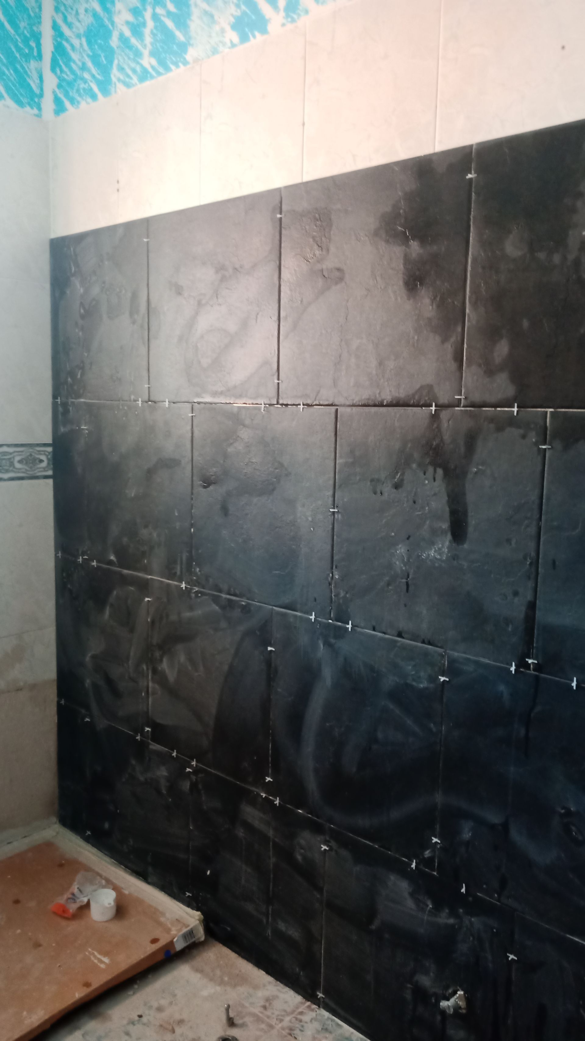 Parede revestida com azulejos pretos em construção em um banheiro, com rejunte e papel de parede azul.