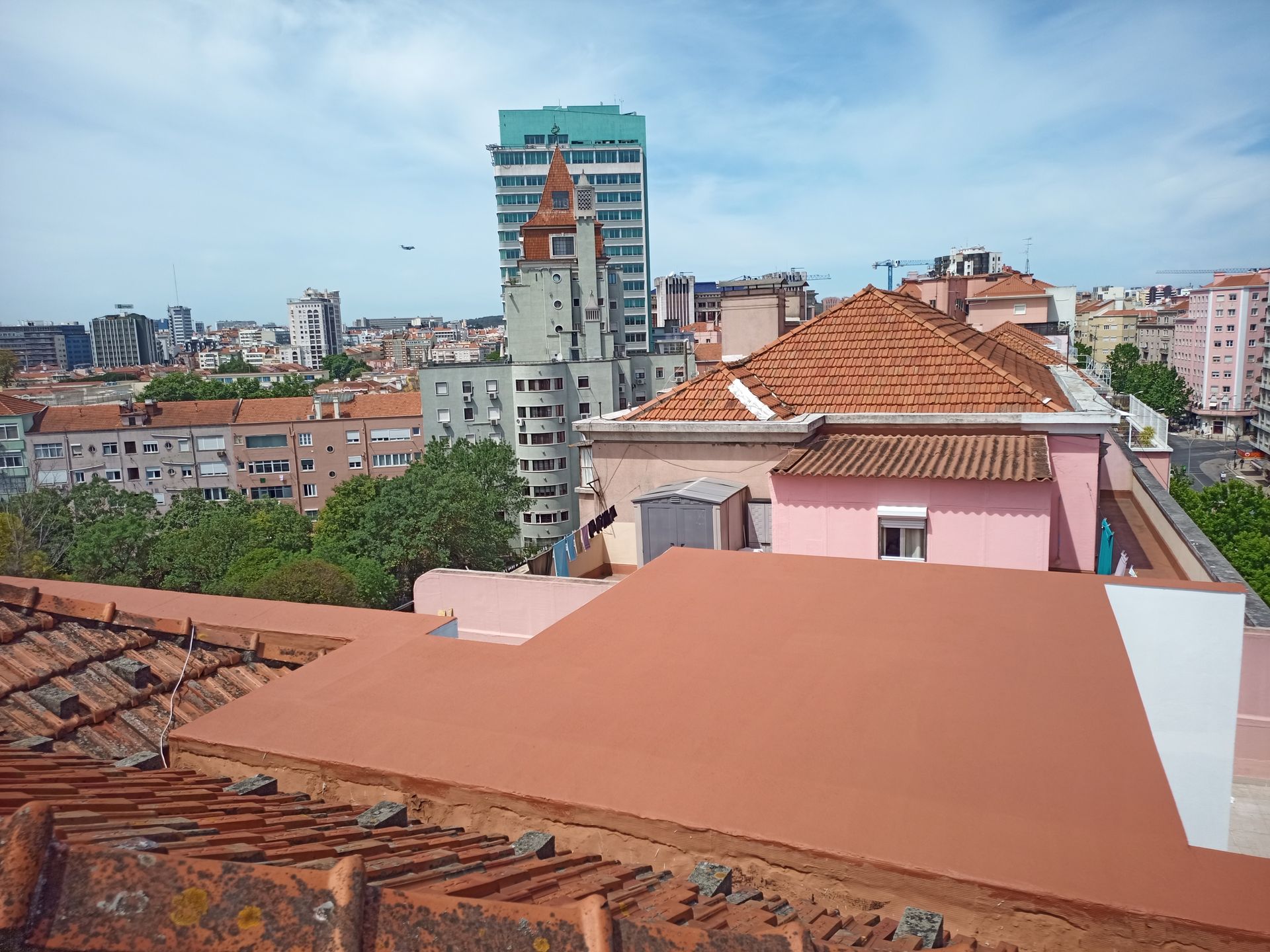 Paisagem urbana com telhados de telha vermelha, edifícios, árvores e céu azul, com um prédio alto e moderno ao fundo.