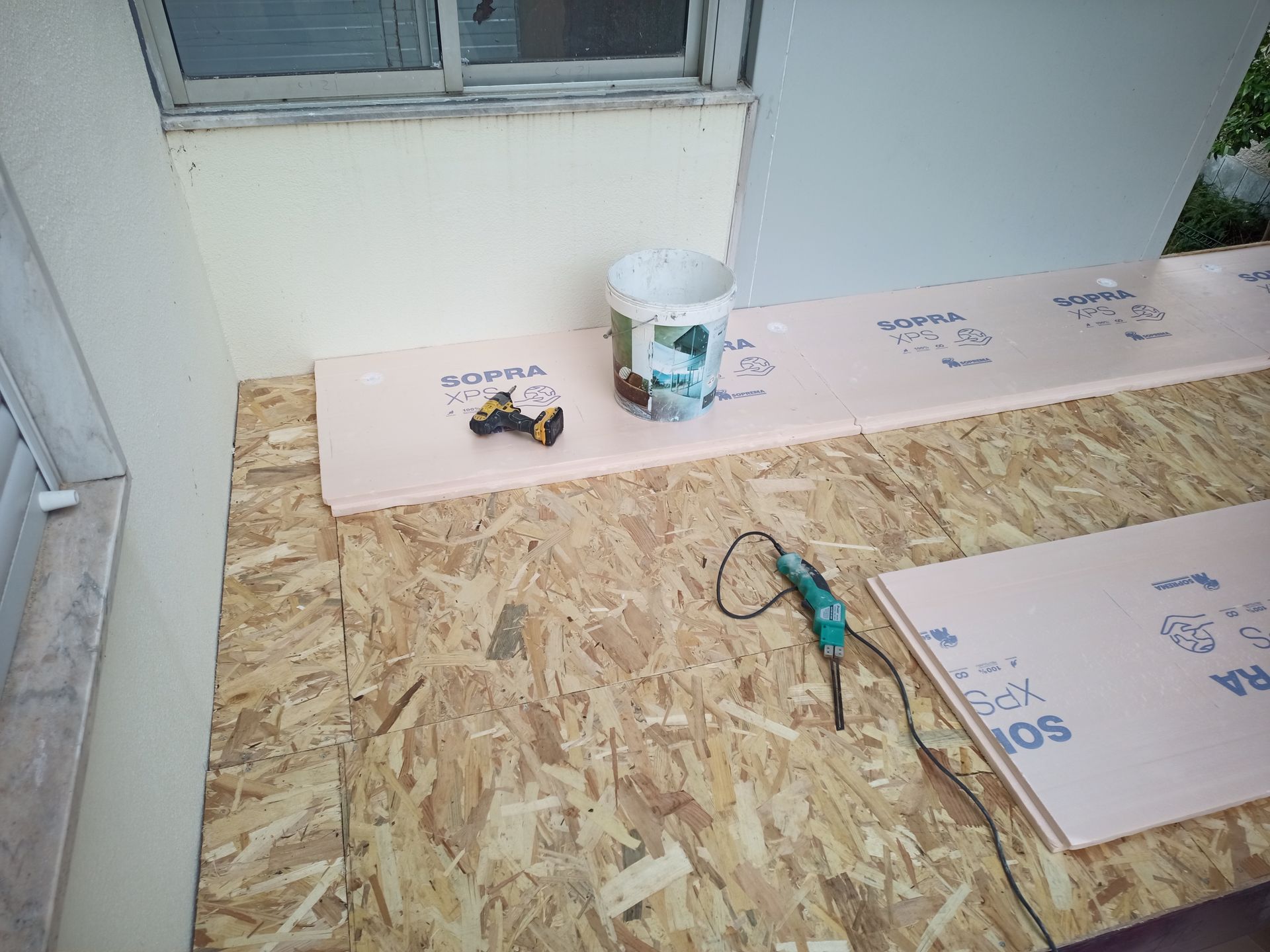 Cena de construção: placas de isolamento colocadas sobre OSB. Um balde, uma furadeira e um cortador de espuma são visíveis na superfície.