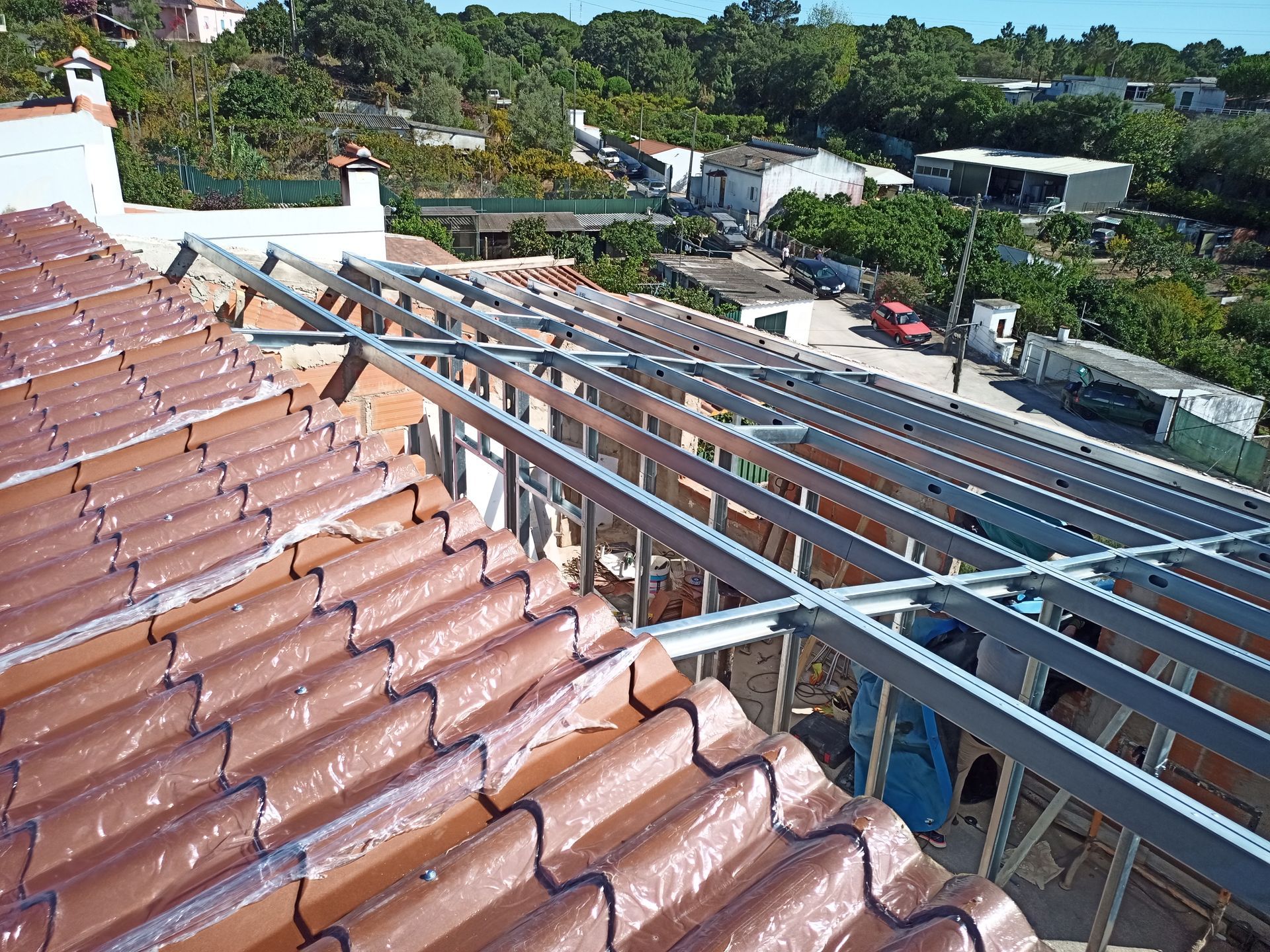Telhado com telhas vermelhas, estrutura metálica parcialmente instalada, ambiente residencial ao fundo.