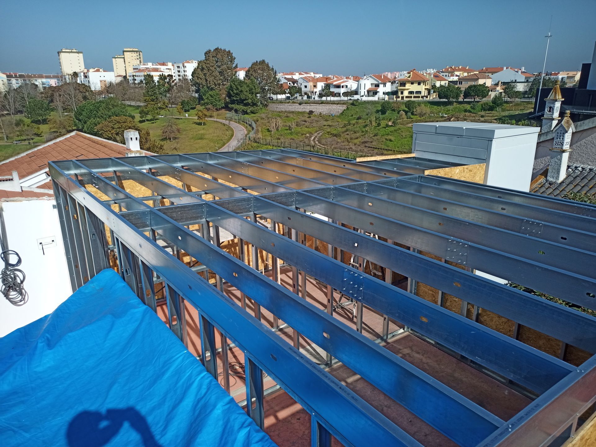 Estrutura metálica para um telhado em construção. Ambiente externo ensolarado com cidade e parque ao fundo.
