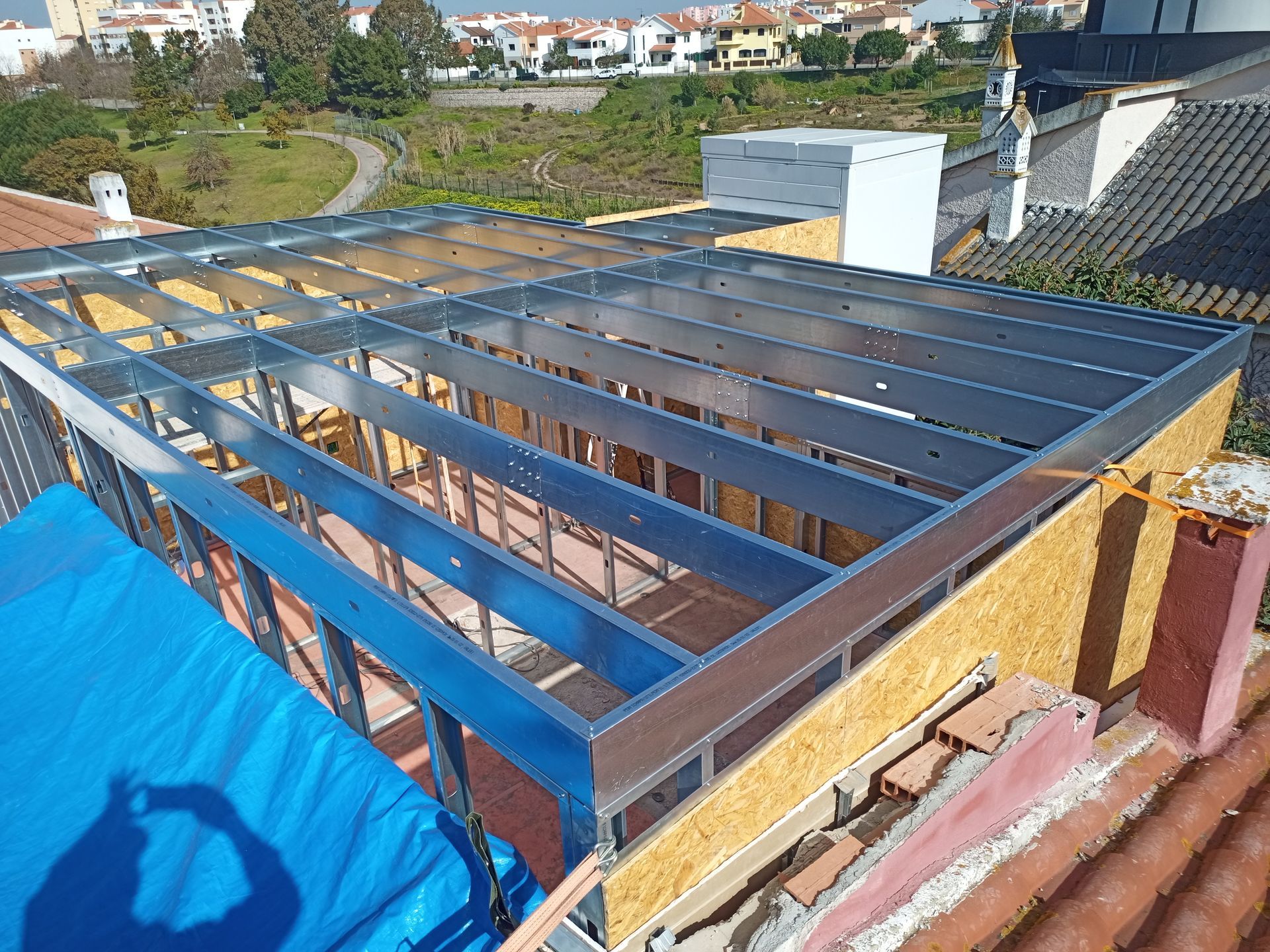 Estrutura metálica sobre o telhado, com revestimento em OSB e lona azul. Prédios ao fundo.