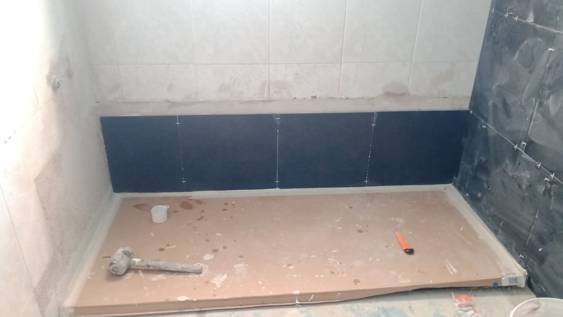 Banheiro em construção, mostrando o trabalho com azulejos. Parede de azulejo preto, base de chuveiro bege, ferramentas presentes.