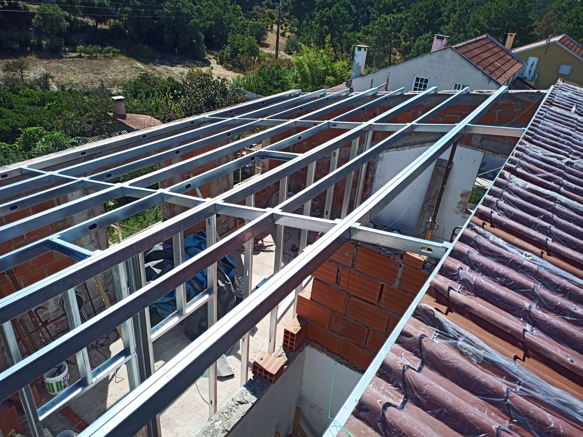 Estrutura metálica sendo construída sobre um telhado de telhas vermelhas, revelando o interior de tijolos e a vegetação ao redor.