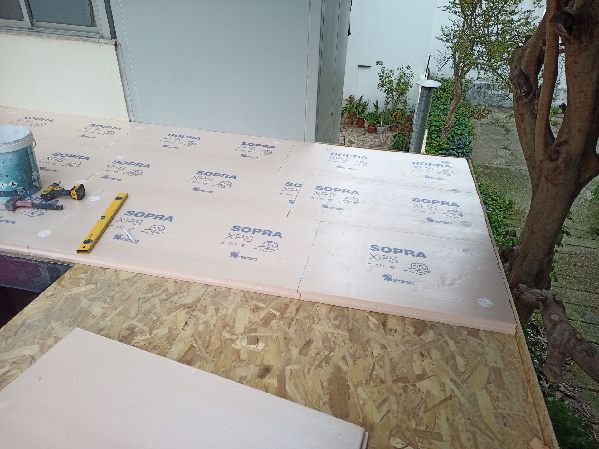 Canteiro de obras: painéis de isolamento de espuma rosa sendo instalados sobre uma placa de OSB; nível e ferramentas visíveis.