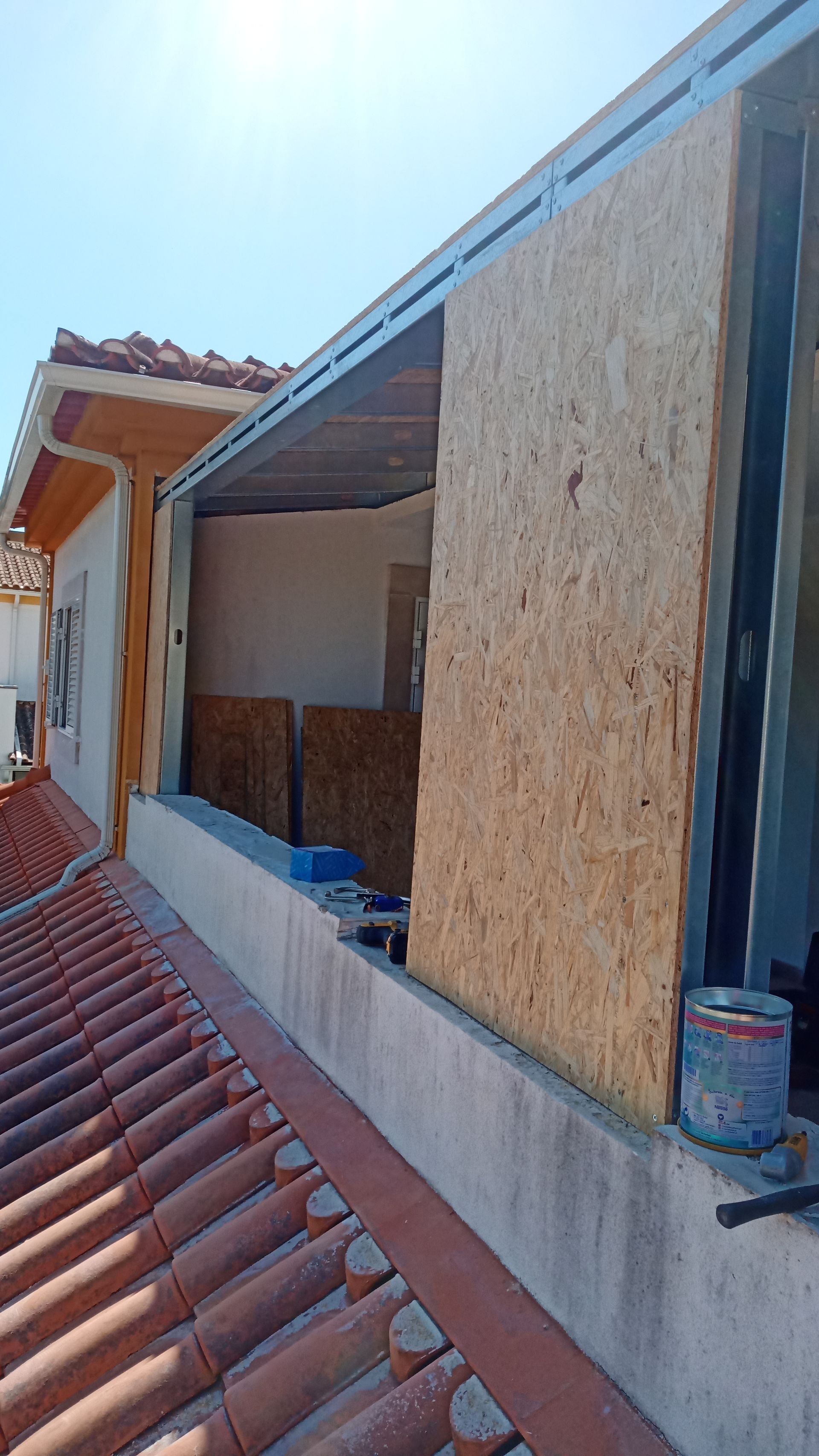 Construção de uma extensão de telhado com painéis de madeira aparentes e estrutura metálica. Dia ensolarado.