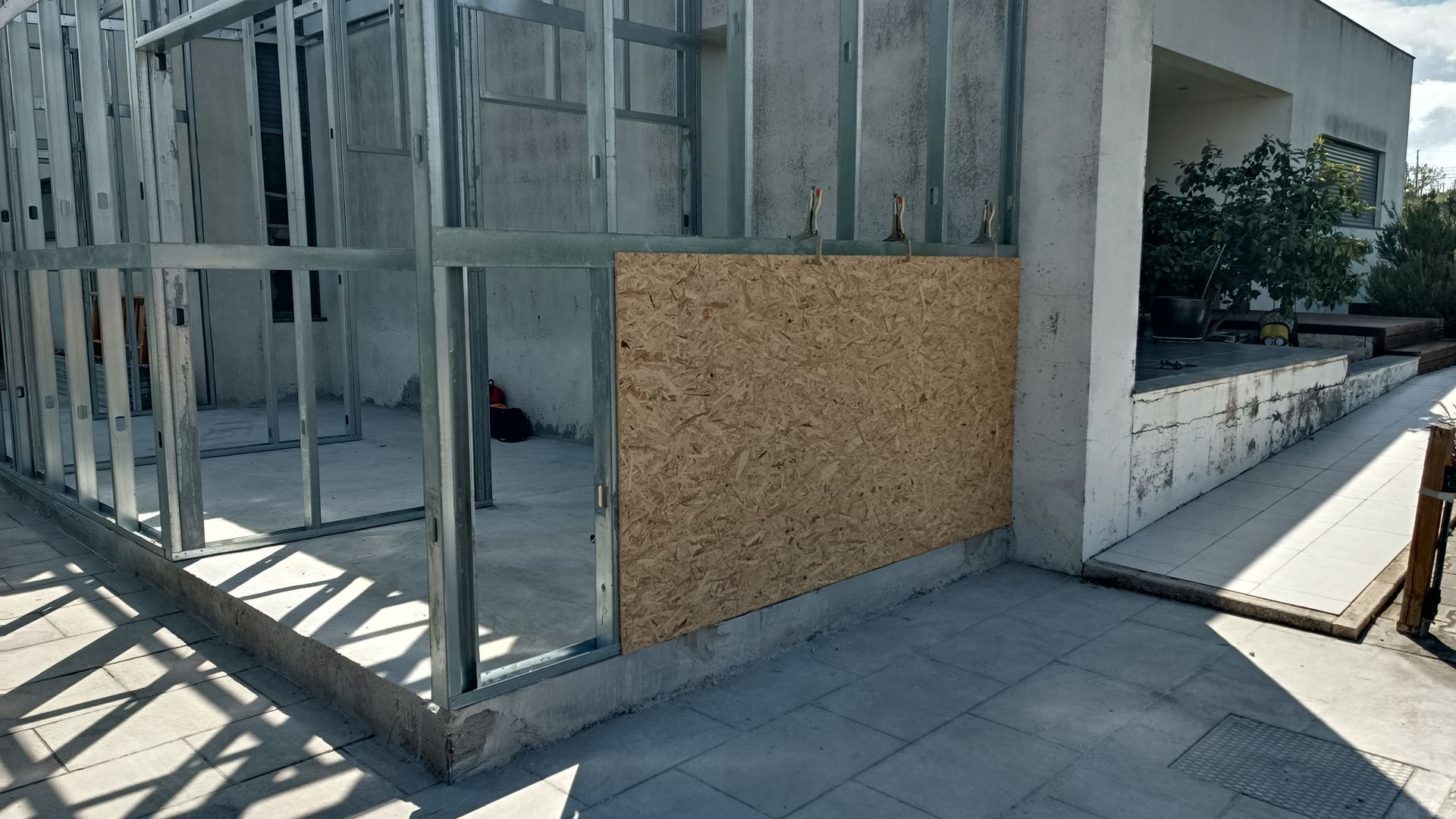 Estrutura de aço para a parede de um edifício, com um painel parcial de madeira fixado, em área externa sobre piso cerâmico.
