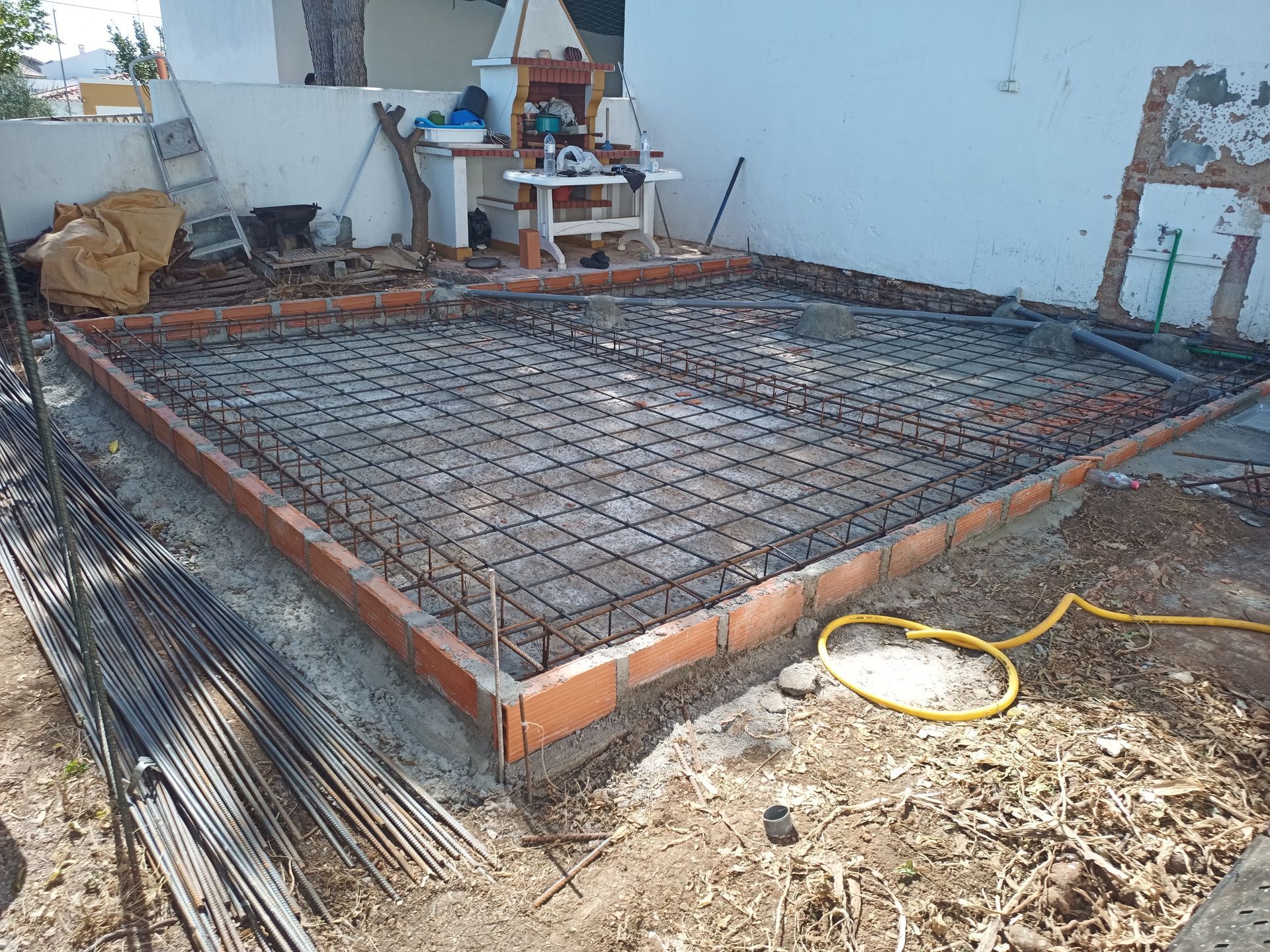 Canteiro de obras com estrutura retangular de tijolos e malha de vergalhões, pronto para a concretagem ao ar livre.