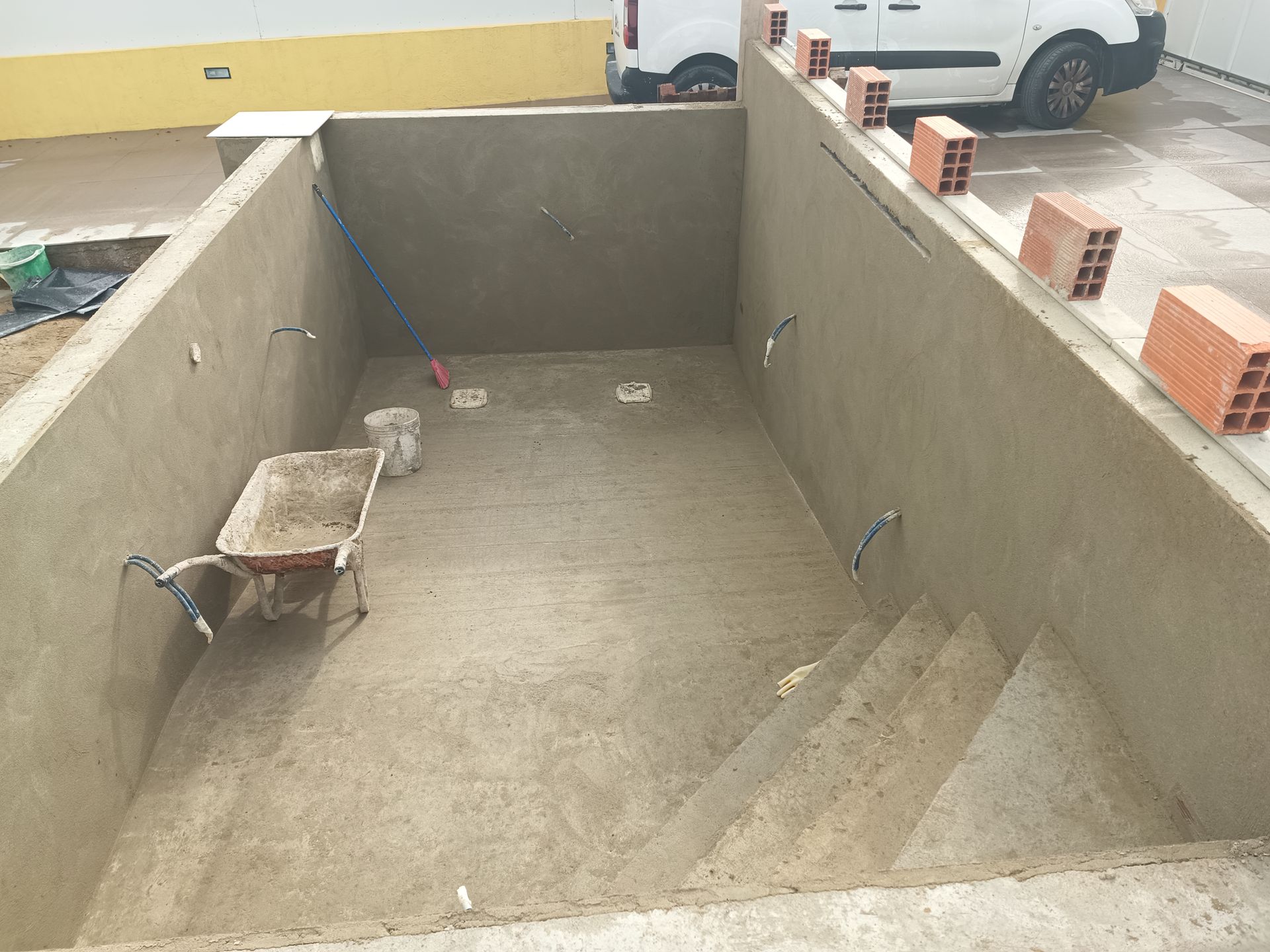 Construção de uma piscina de concreto. Paredes e piso cinza, degraus, carrinho de mão e canos de água visíveis.