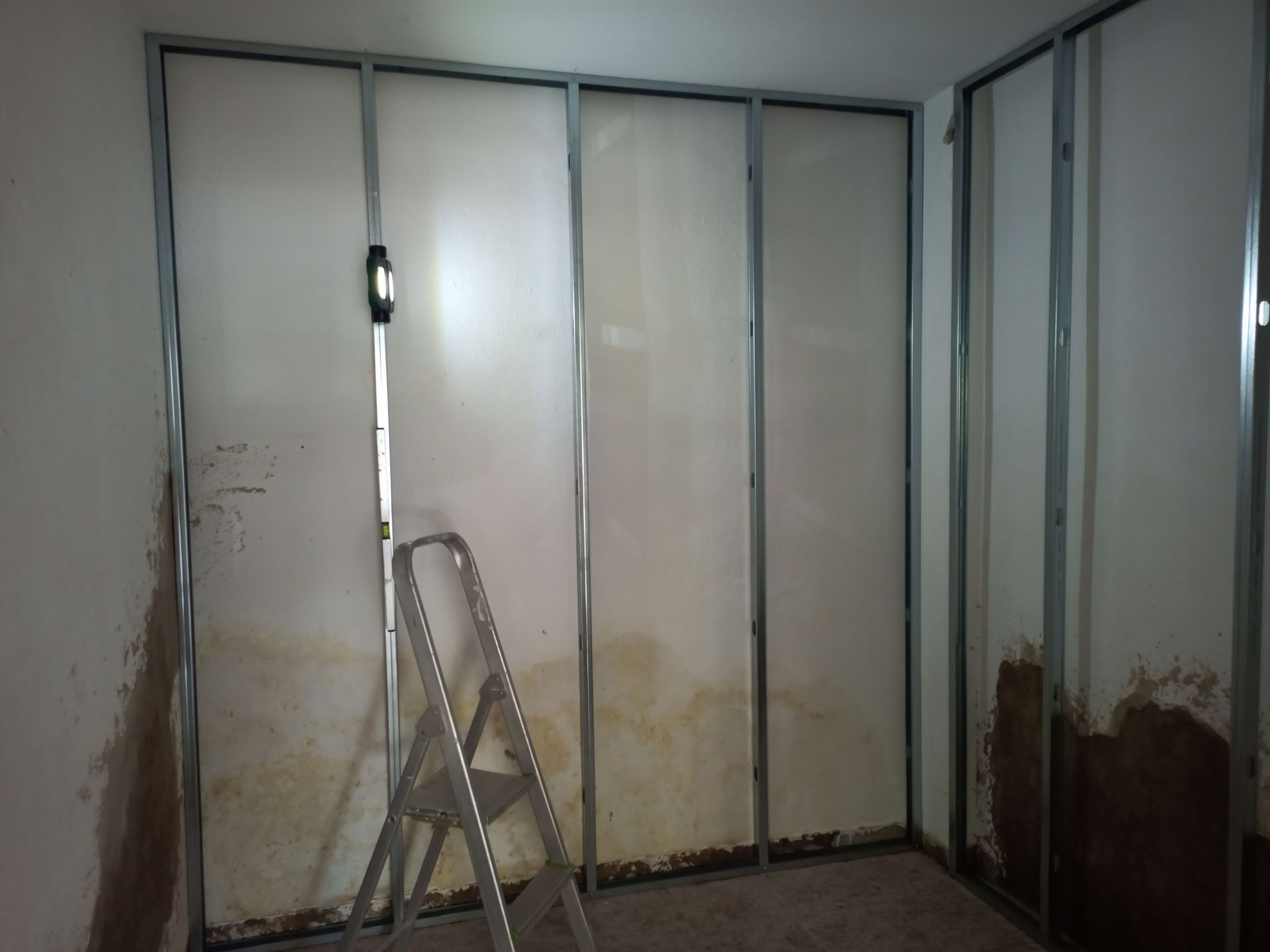 Quarto interior com estrutura de metal e paredes danificadas; uma escada de mão está encostada na parede.