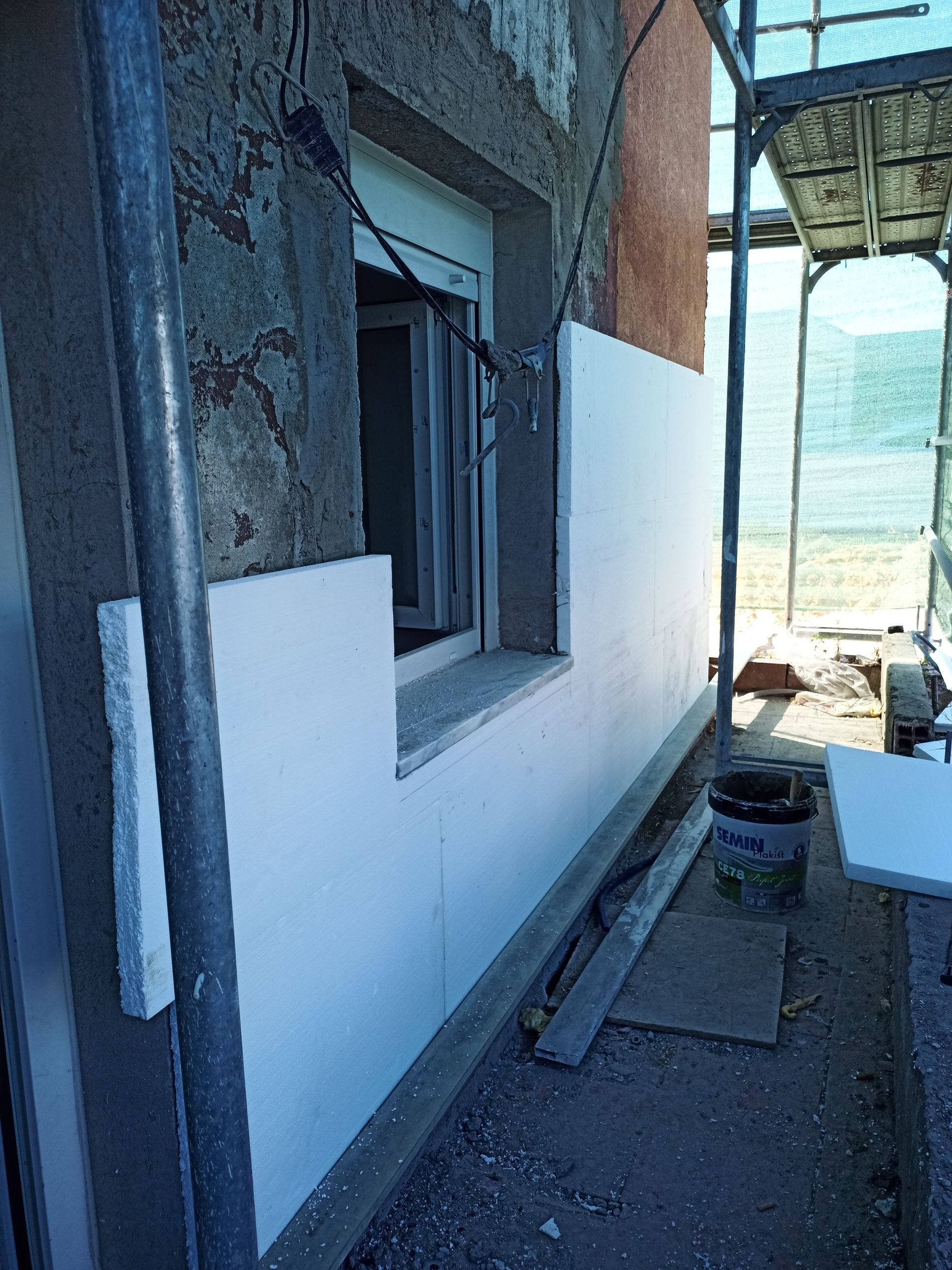 Parede externa com painéis isolantes brancos sendo instalados ao redor de uma janela. Há andaimes no local.