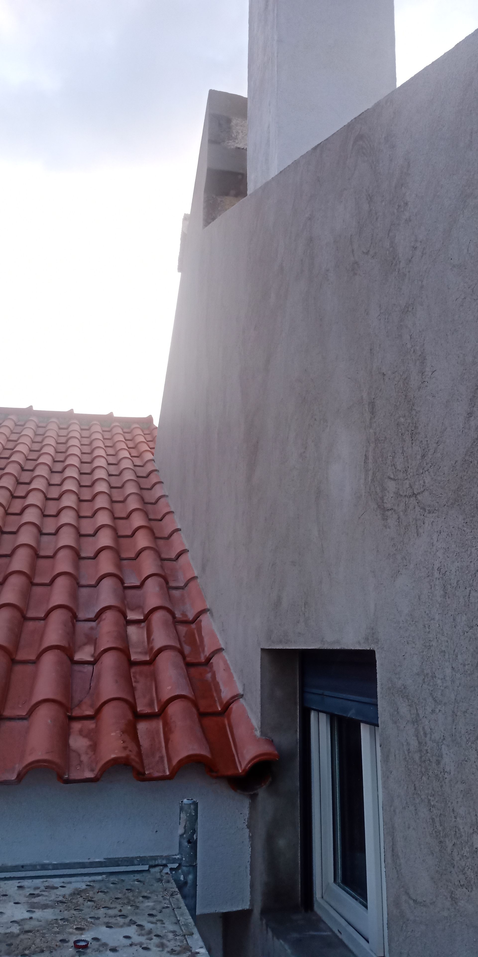 Telhado de telha vermelha ao lado de um prédio cinza claro com janela, chaminé e céu nublado.